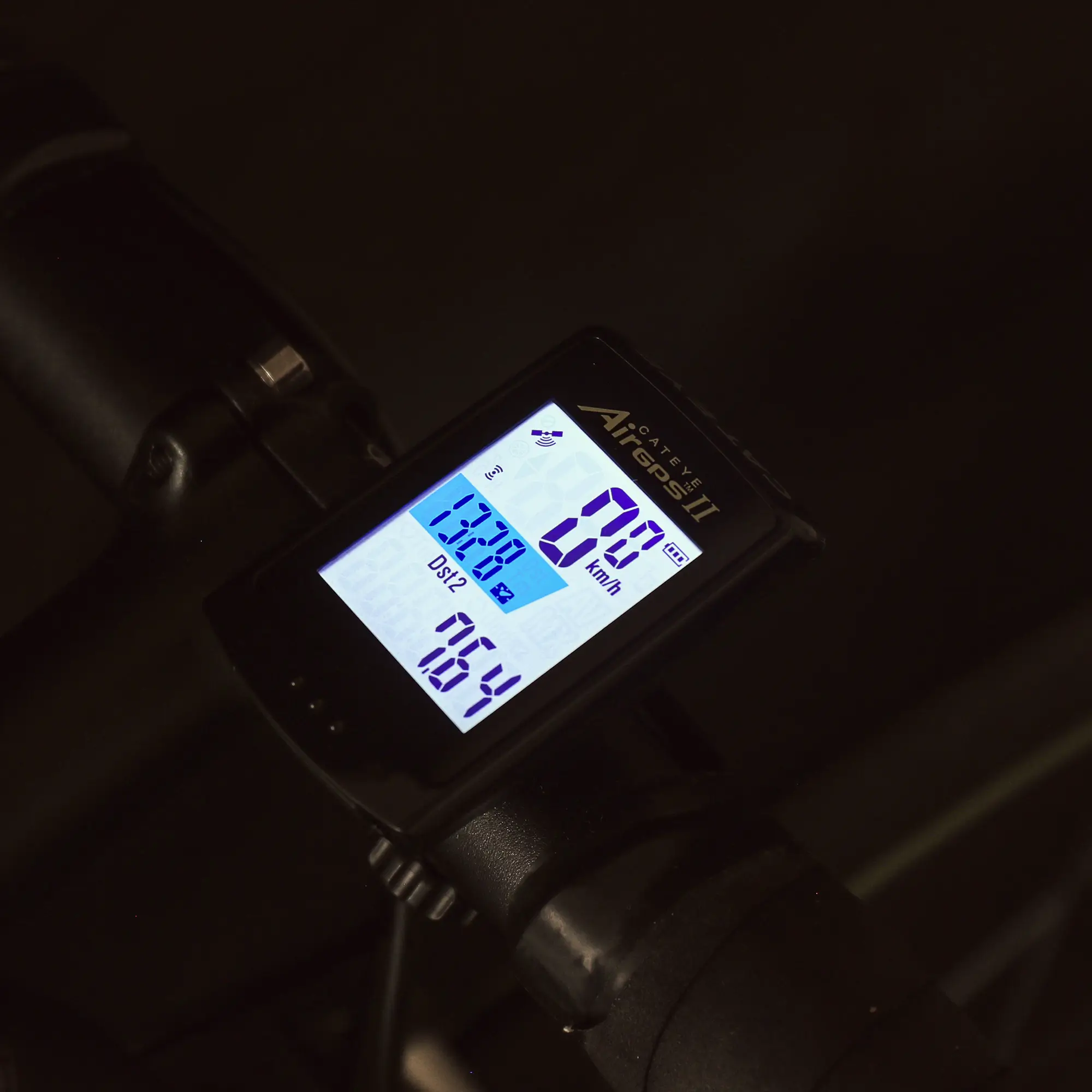 A close-up of the CATEYE AirGPS II digital bike computer on handlebars, showing speed (km/h), ride time, distance, and Strava integration on a blue and white backlit display.