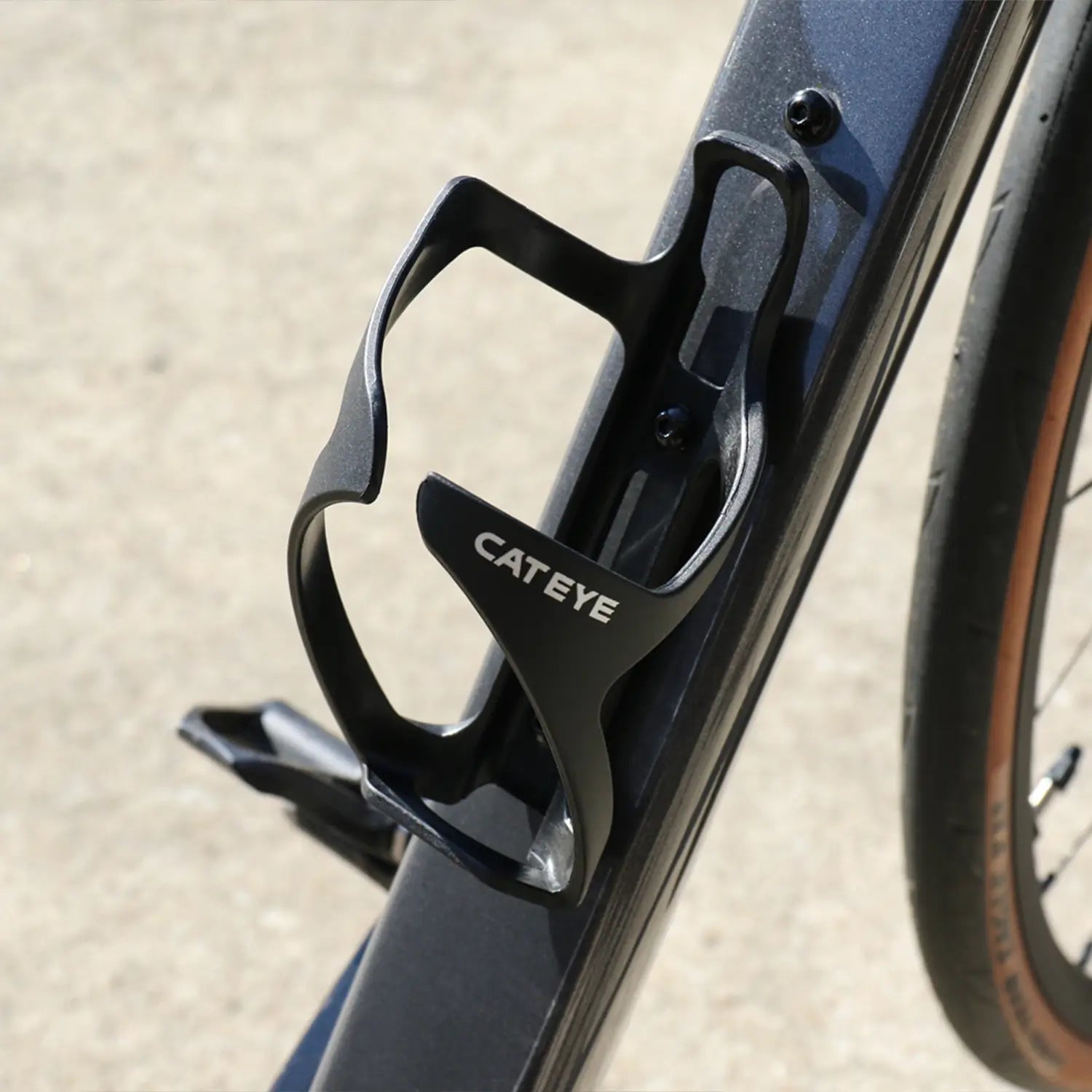 A close-up of a CATEYE BC-500 side-loading bottle cage in black, mounted on the bike frame—ideal for bikepacking—with part of the rear wheel visible in the background.