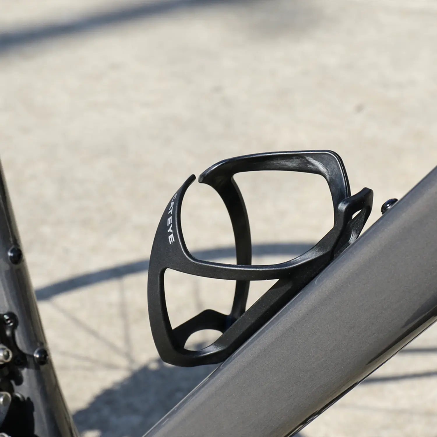 A close-up of the CATEYE BC-500 Bottle Cage, a black side-loading model mounted on a gray bike frame outdoors on a sunny day—perfect for bikepacking adventures.