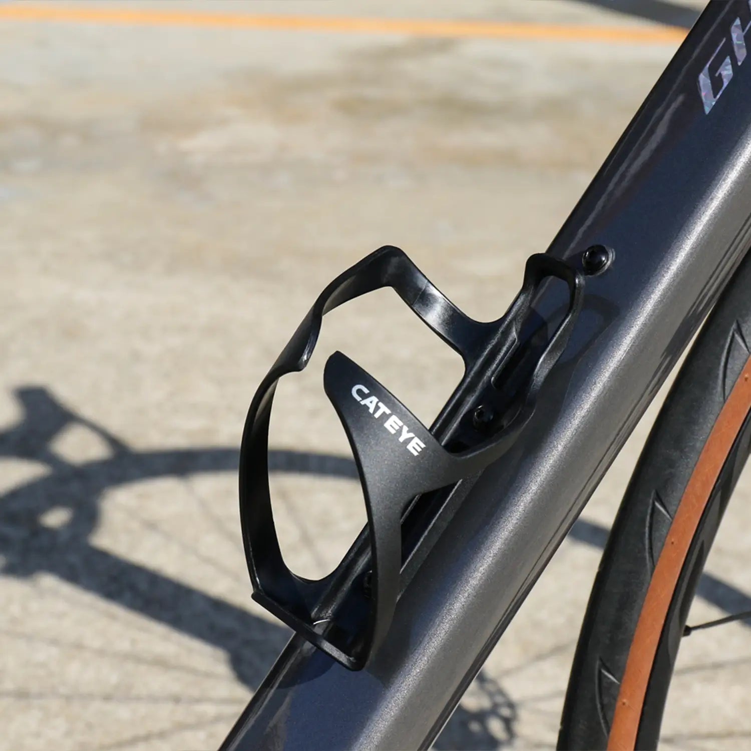 A close-up of a CATEYE BC-500 Bottle Cage in black, mounted on a bike frame with the wheel and tire visible on sunlit pavement—ideal for quick access to your bottle during bikepacking adventures.