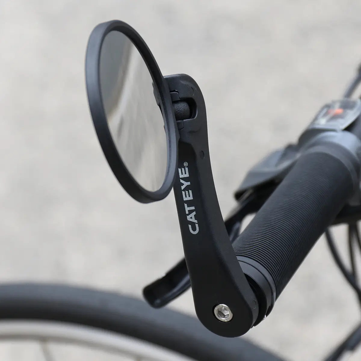 Close-up of a CATEYE BM-600 Handlebar Mirror with a stainless steel lens attached to a black bike handlebar, providing enhanced rear visibility against a blurred pavement background.