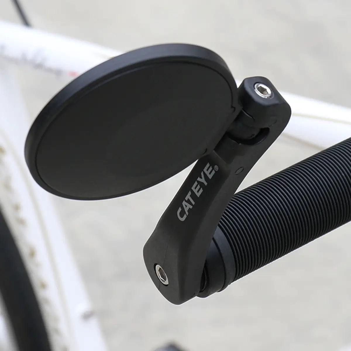 A close-up of a black CATEYE BM-600 Handlebar Mirror with a stainless steel lens, mounted on the handlebar of a white bicycle, enhancing rear visibility against a blurred background.