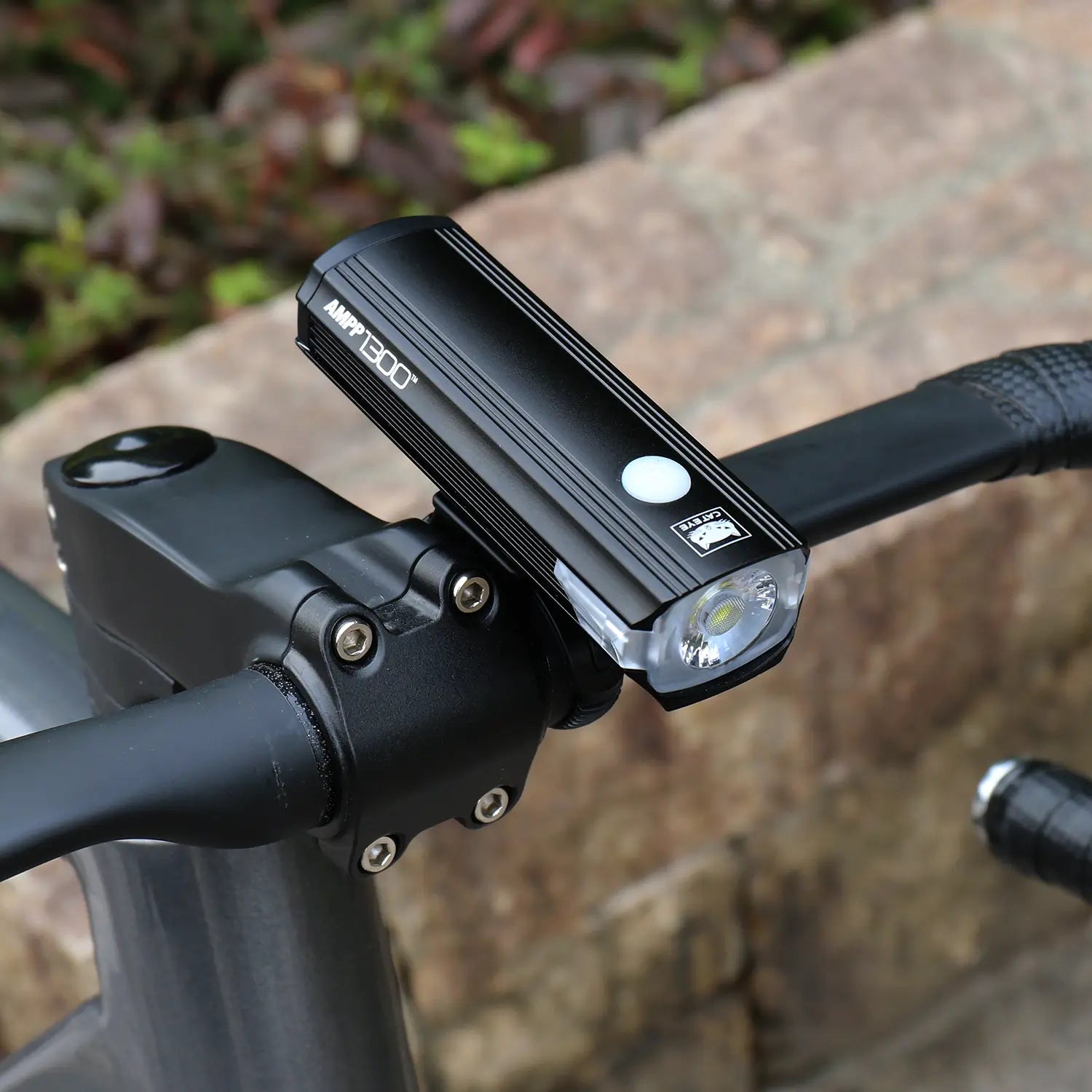 A black CATEYE AMPP1300 bicycle headlight with OptiCube lens technology is mounted on bike handlebars, with a stone wall and foliage blurred in the background.