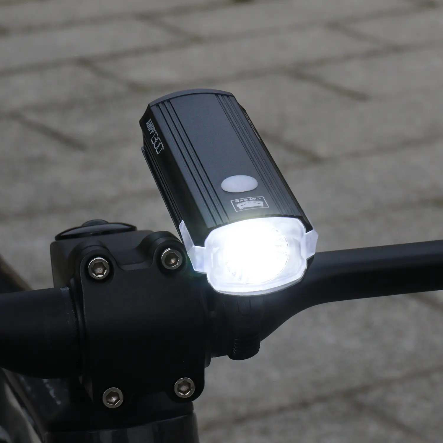 A CATEYE AMPP1300 LED bicycle headlight with OptiCube lens technology is mounted on a black handlebar, casting a white beam, while gray paving stones form a softly blurred background.