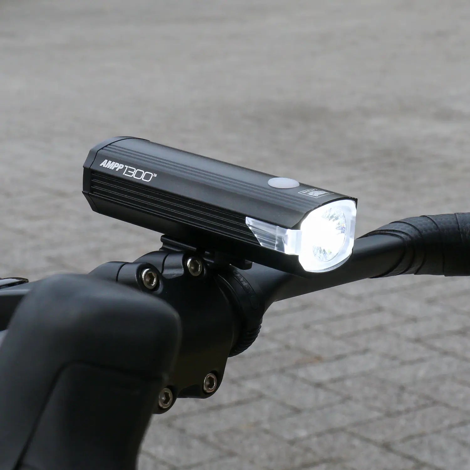 A black CATEYE AMPP1300 bicycle headlight with OptiCube lens technology is mounted on the handlebars, turned on and shining brightly against an outdoor paved background.