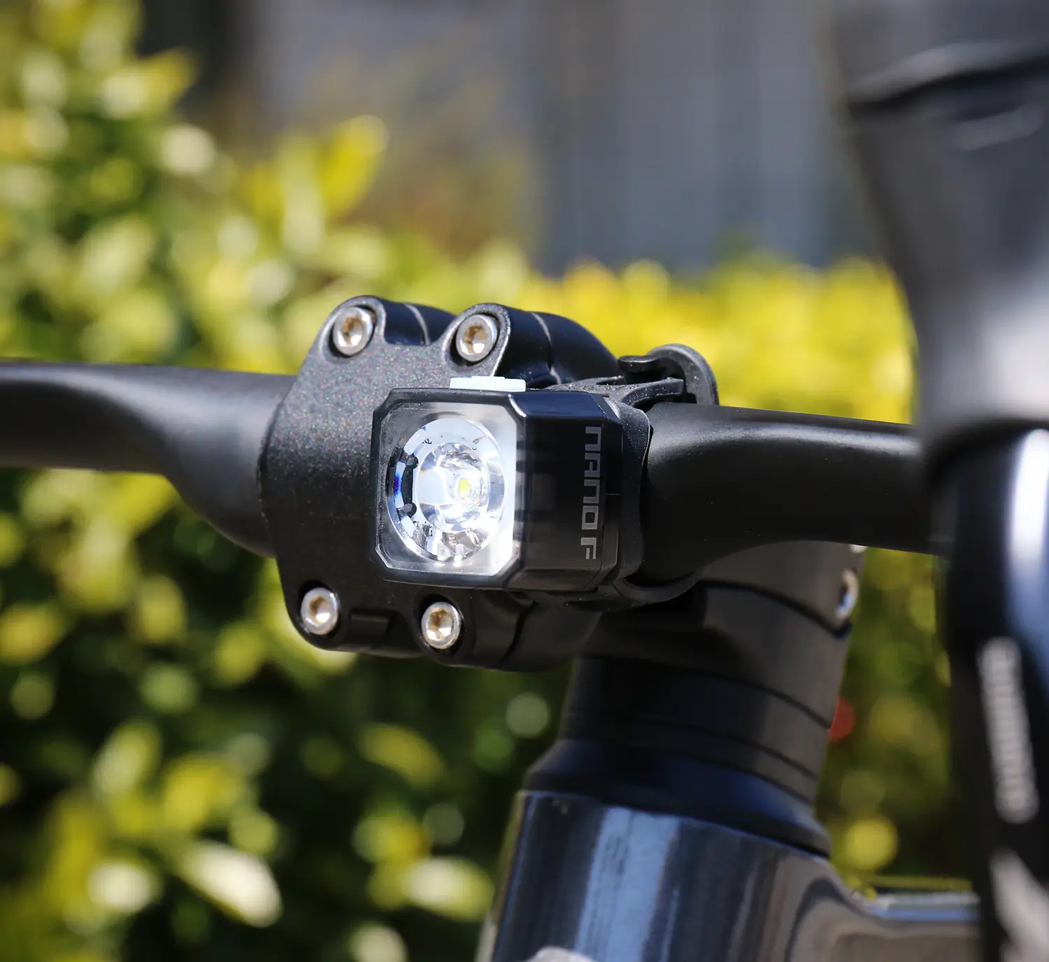 Close-up of a black bicycle handlebar with a CATEYE NANO100 F daytime front safety light mounted and turned on, emitting a 100-lumen beam against a blurred green foliage background.