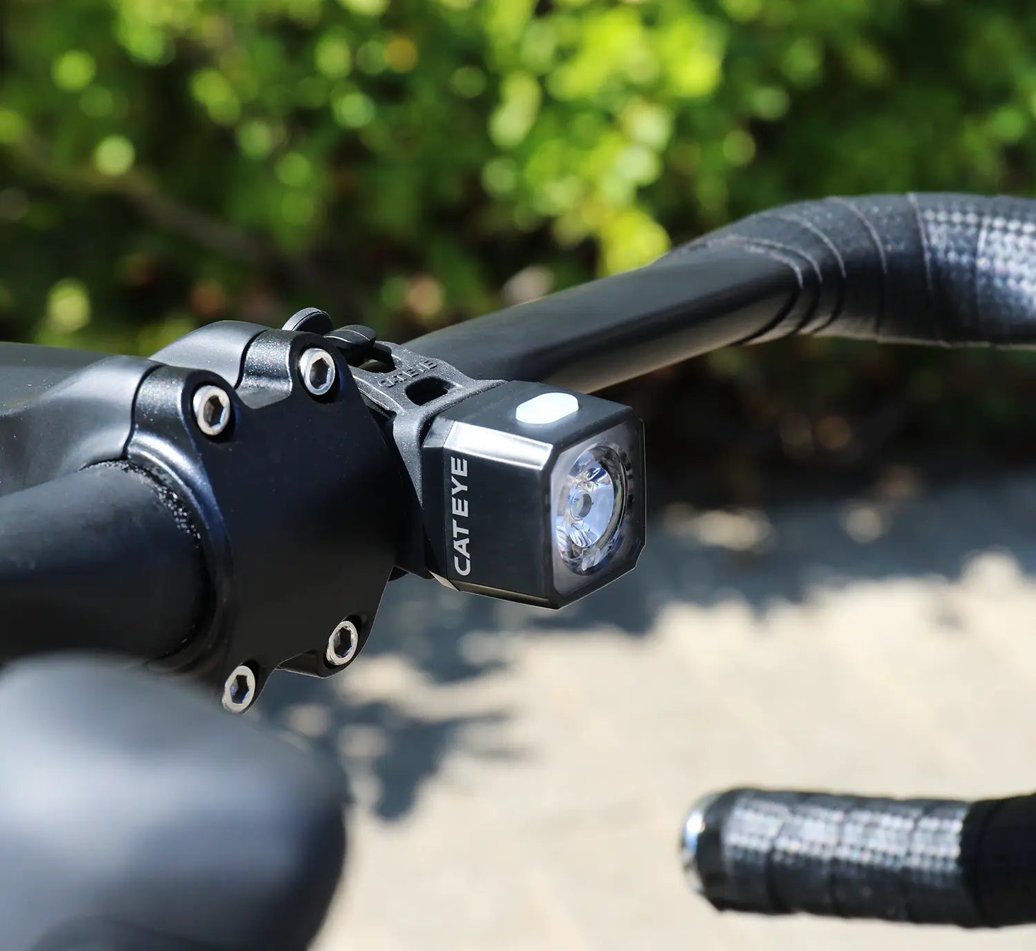 A close-up of a black bicycle handlebar shows the CATEYE NANO100 F, a USB-C rechargeable bike light with a 100-lumen beam, mounted against a blurred green background.