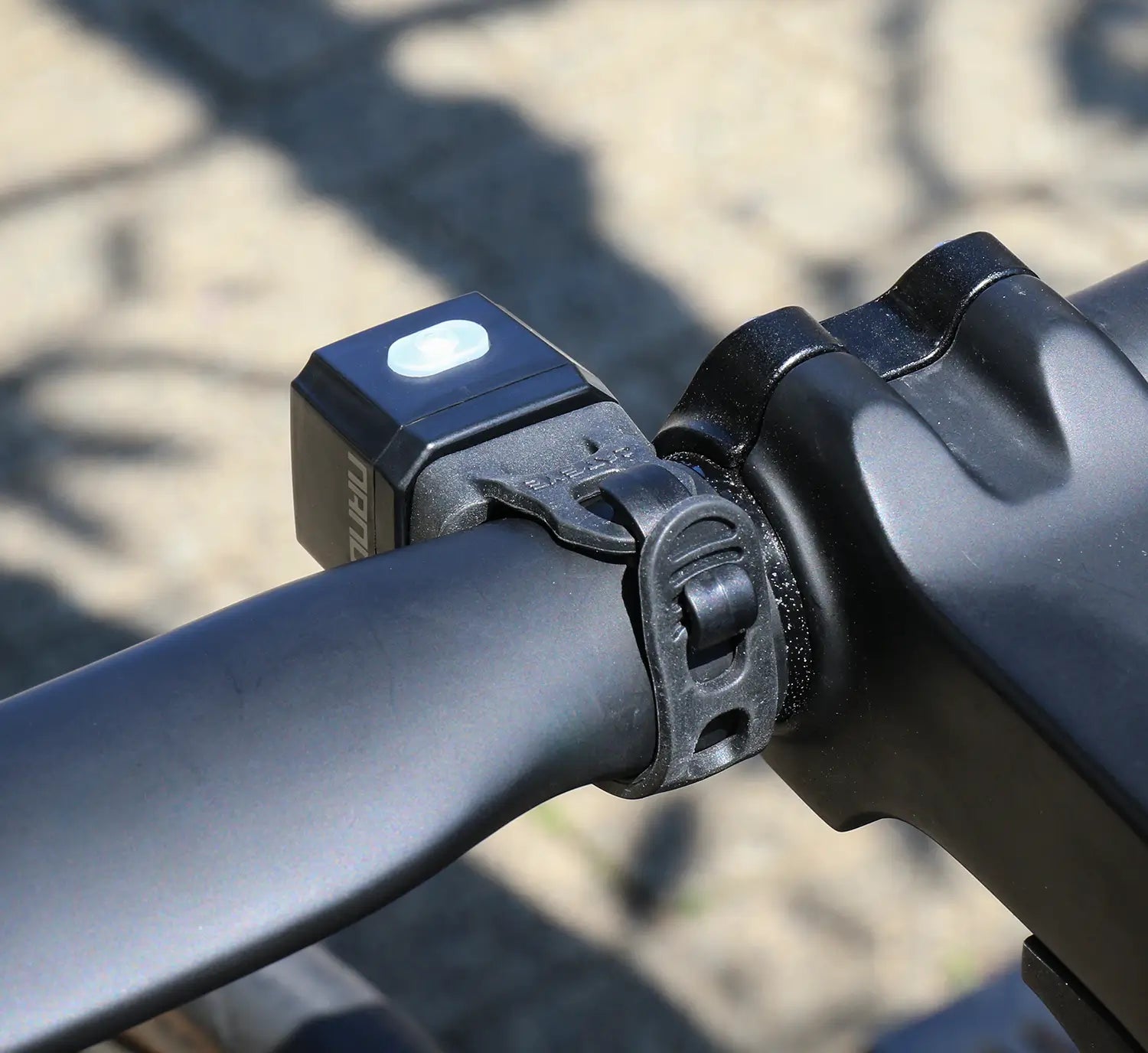 Close-up of a bicycle handlebar featuring the CATEYE NANO100 F, a USB-C rechargeable bike light with a white button, secured with a black rubber mount near the stem. The background is blurred.