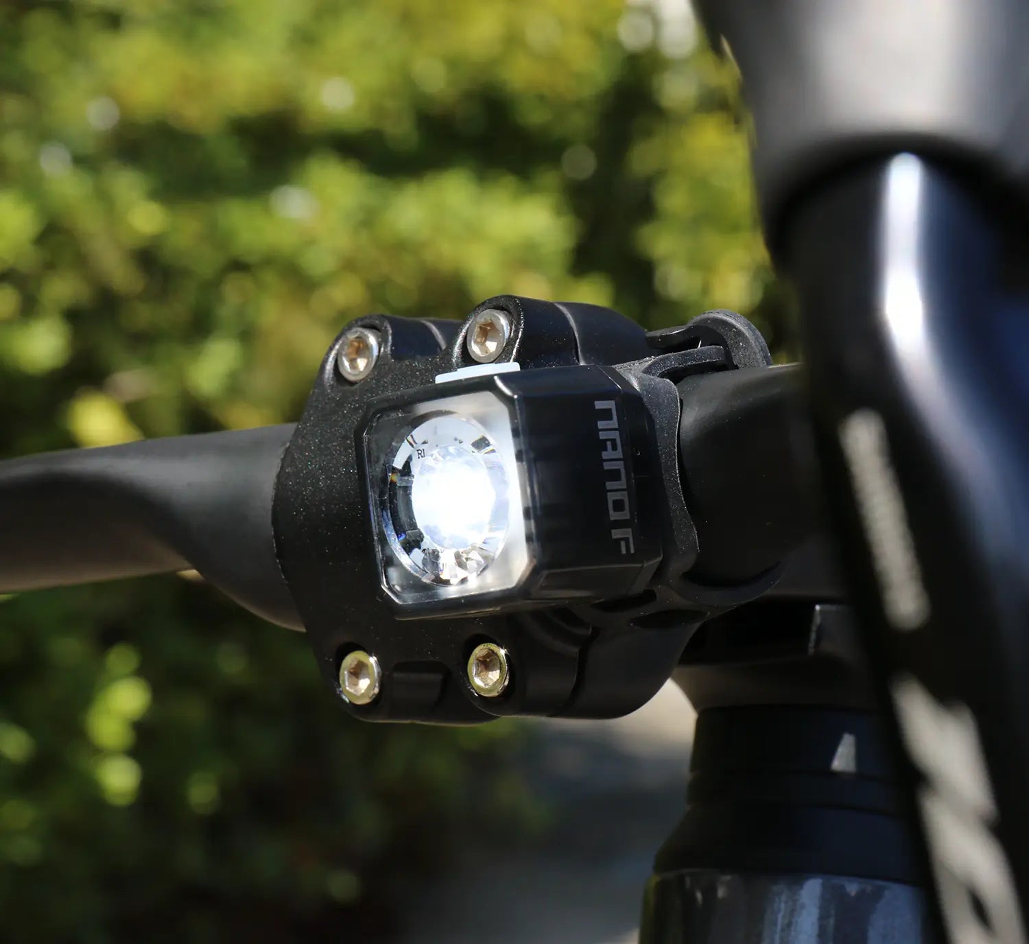 Close-up of the CATEYE NANO100 F, a USB-C rechargeable bike light with a 100-lumen beam, mounted on a black handlebar against a softly blurred green background.