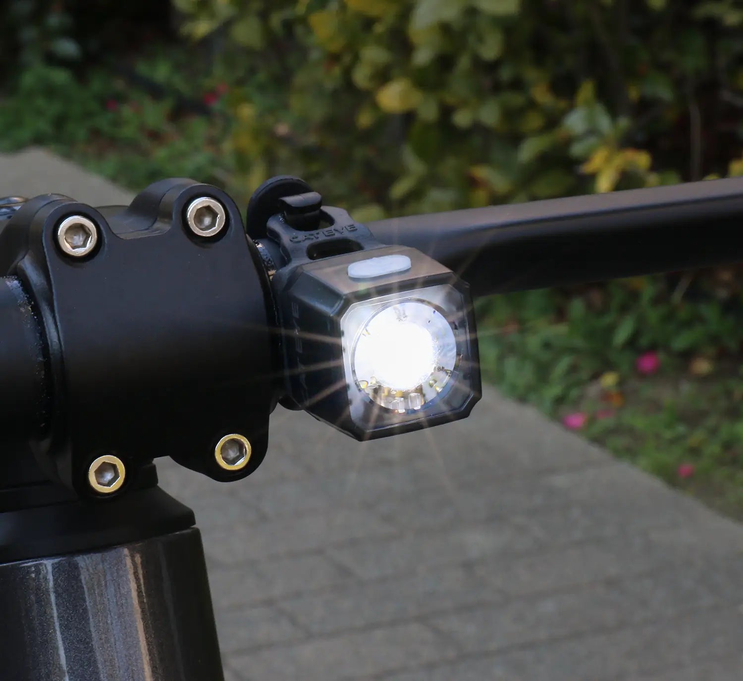 The CATEYE NANO100 F, a USB-C rechargeable bike light, is mounted on the handlebars and shines a bright 100-lumen white beam outdoors with greenery and a paved path in the background.