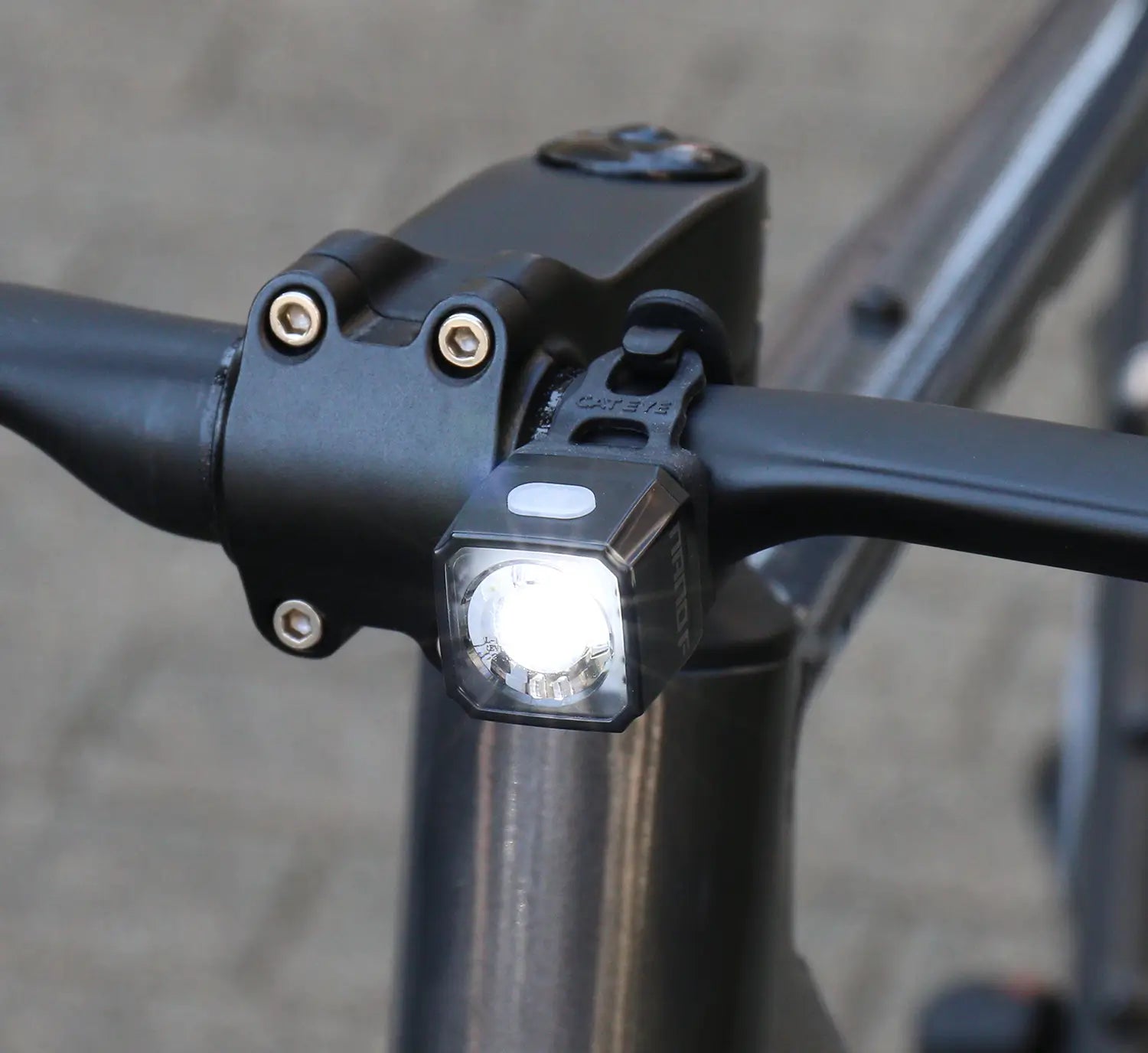 A close-up of the CATEYE NANO100 F USB-C rechargeable bike light, mounted by the handlebar stem and turned on, casts a 100-lumen beam onto blurred pavement in the background.