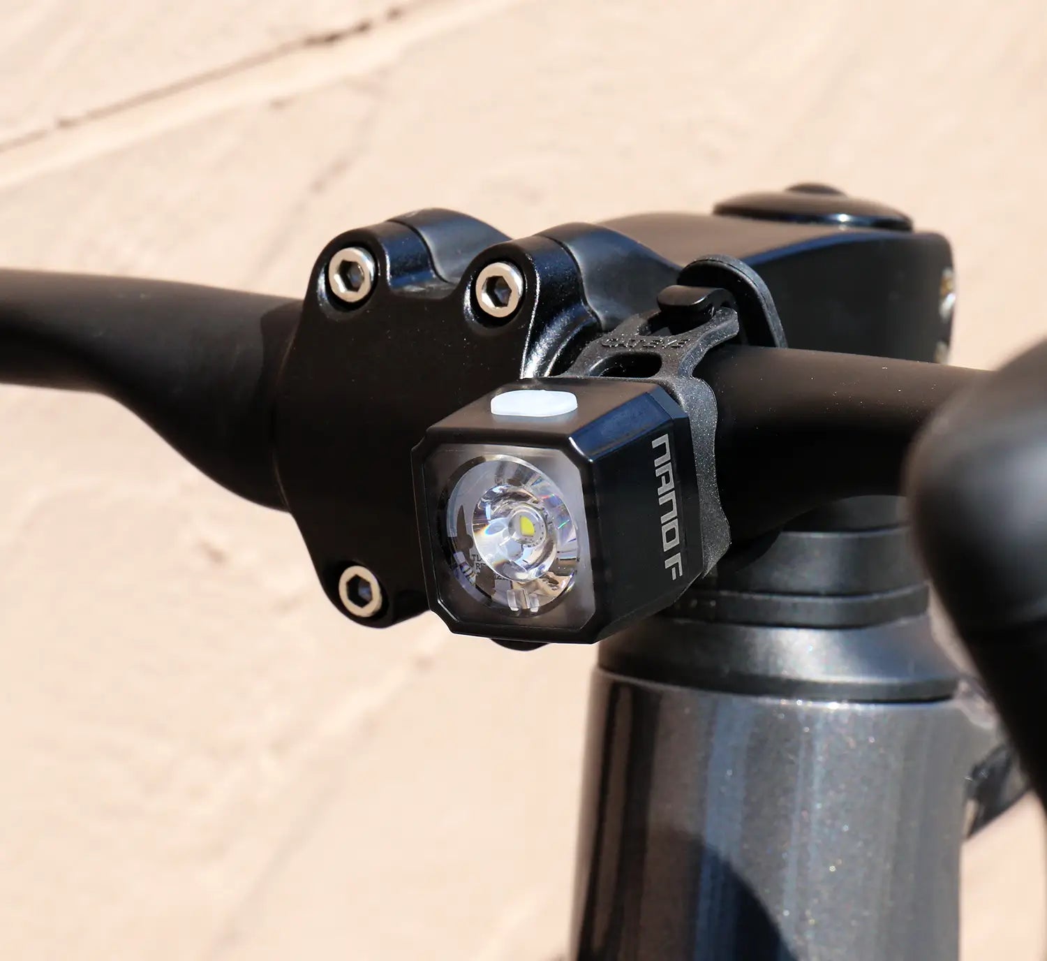 Close-up of a bicycle handlebar featuring the CATEYE NANO100 F, a USB-C rechargeable bike light. The 100-lumen beam provides visibility, while a textured, light-colored wall serves as the background.