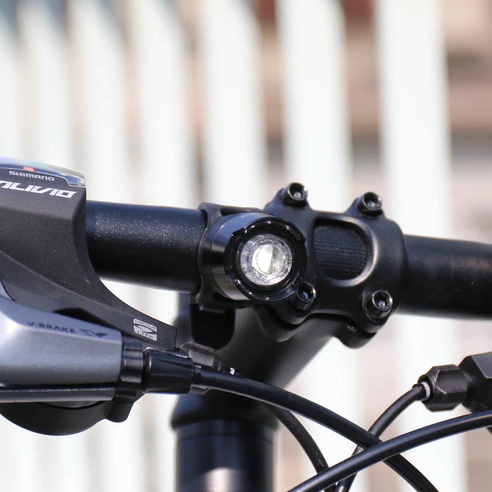Close-up of a CATEYE ORB Rechargeable bike light mounted on a bicycle handlebar with gear shifter, brake lever, and cables, set against a blurred background of vertical white bars.