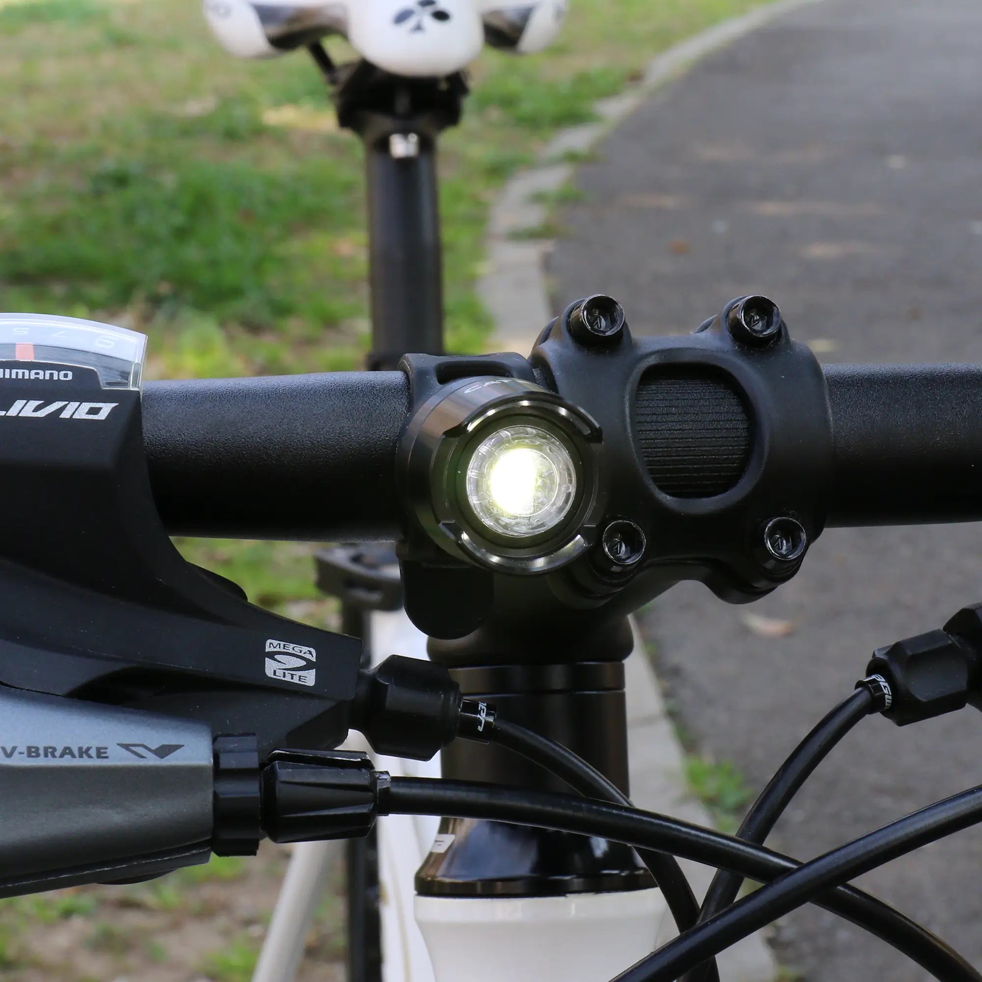 Close-up of a CATEYE ORB Rechargeable LED light mounted on a bicycle handlebar, with brake lever and gear shifter visible; blurred greenery and paved path in the background.