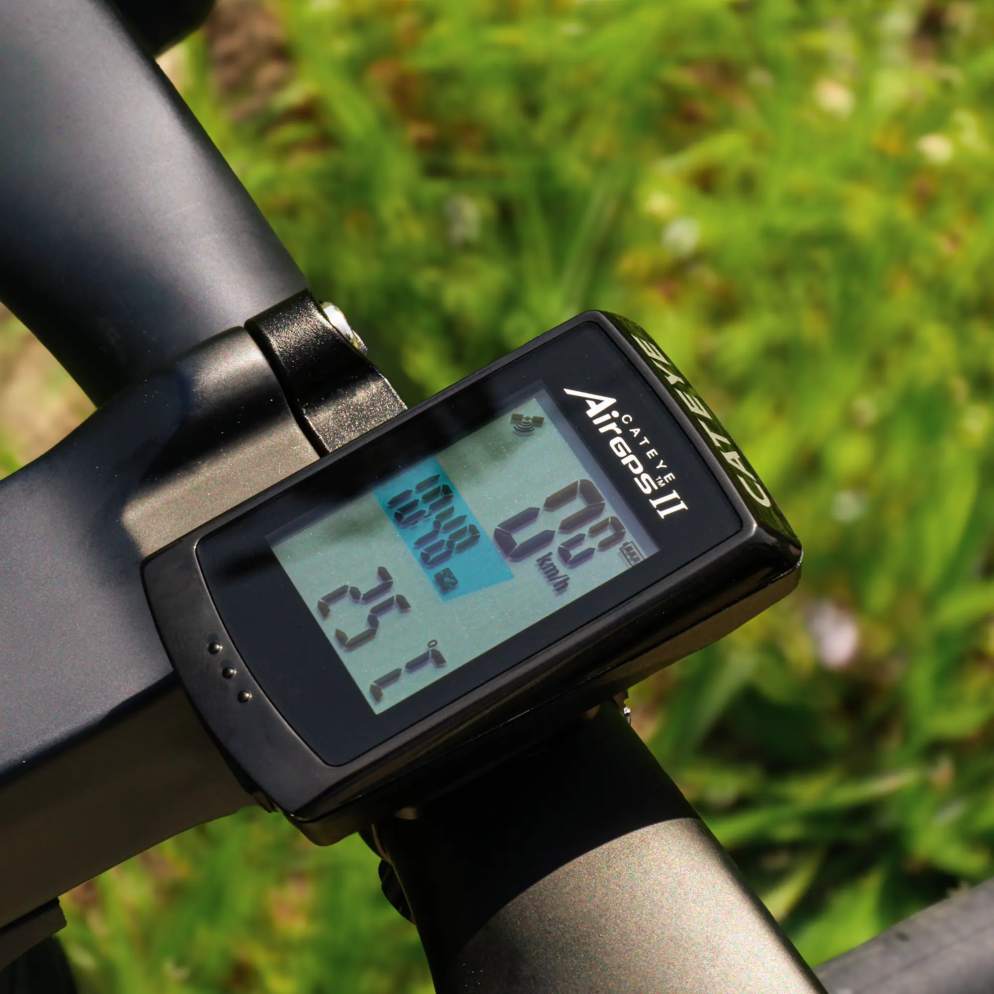 A close-up of a CATEYE AirGPS II bike computer on a handlebar shows speed, distance, and temperature with high GPS accuracy, set against a blurred green grass and plant background.