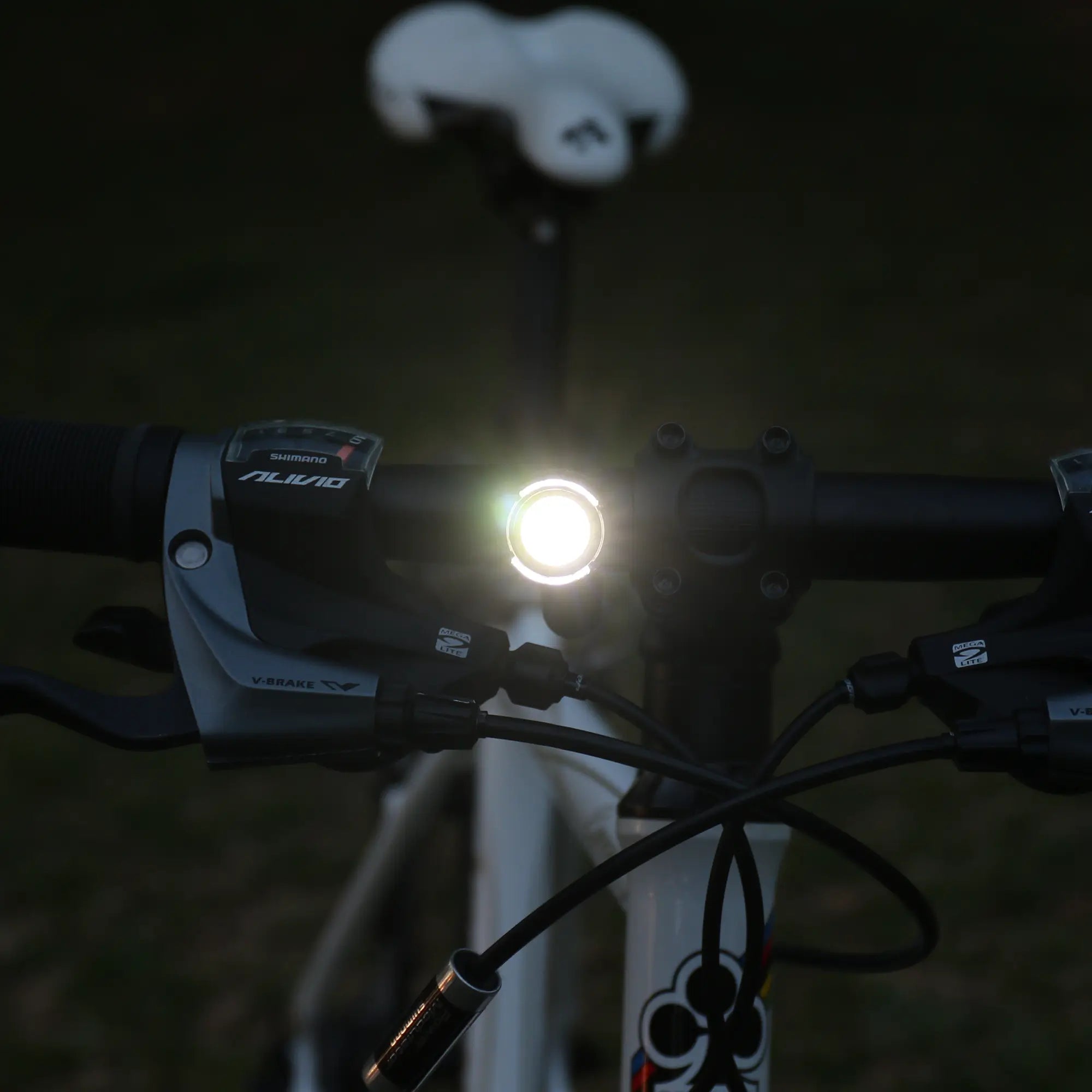 Close-up of a bicycle’s handlebars featuring the CATEYE ORB Rechargeable LED safety light glowing in low light, with a white saddle blurred in the background.