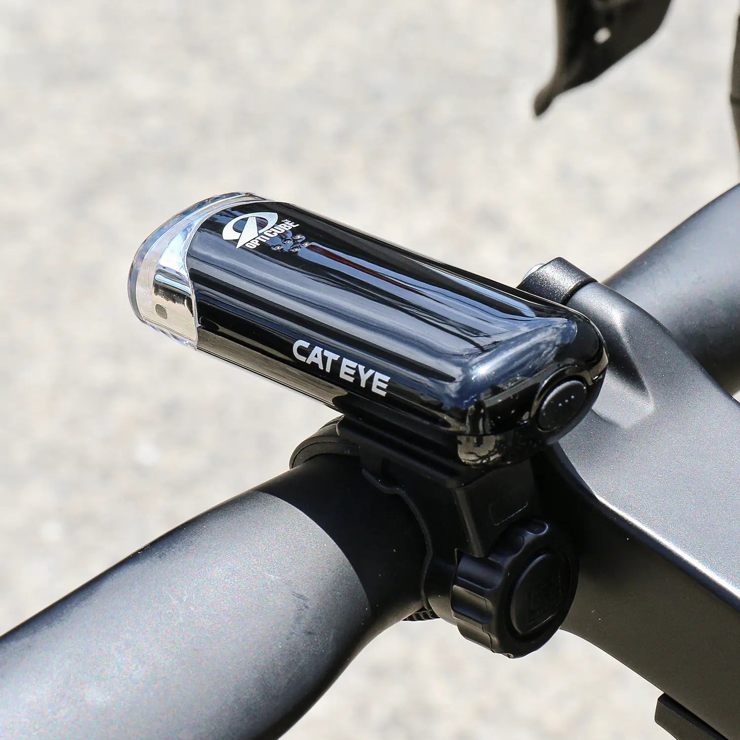 A close-up of a CATEYE Hl-El135N bicycle headlight with OptiCube lens technology, securely mounted on a handlebar with a black clamp, shown outdoors in daylight.