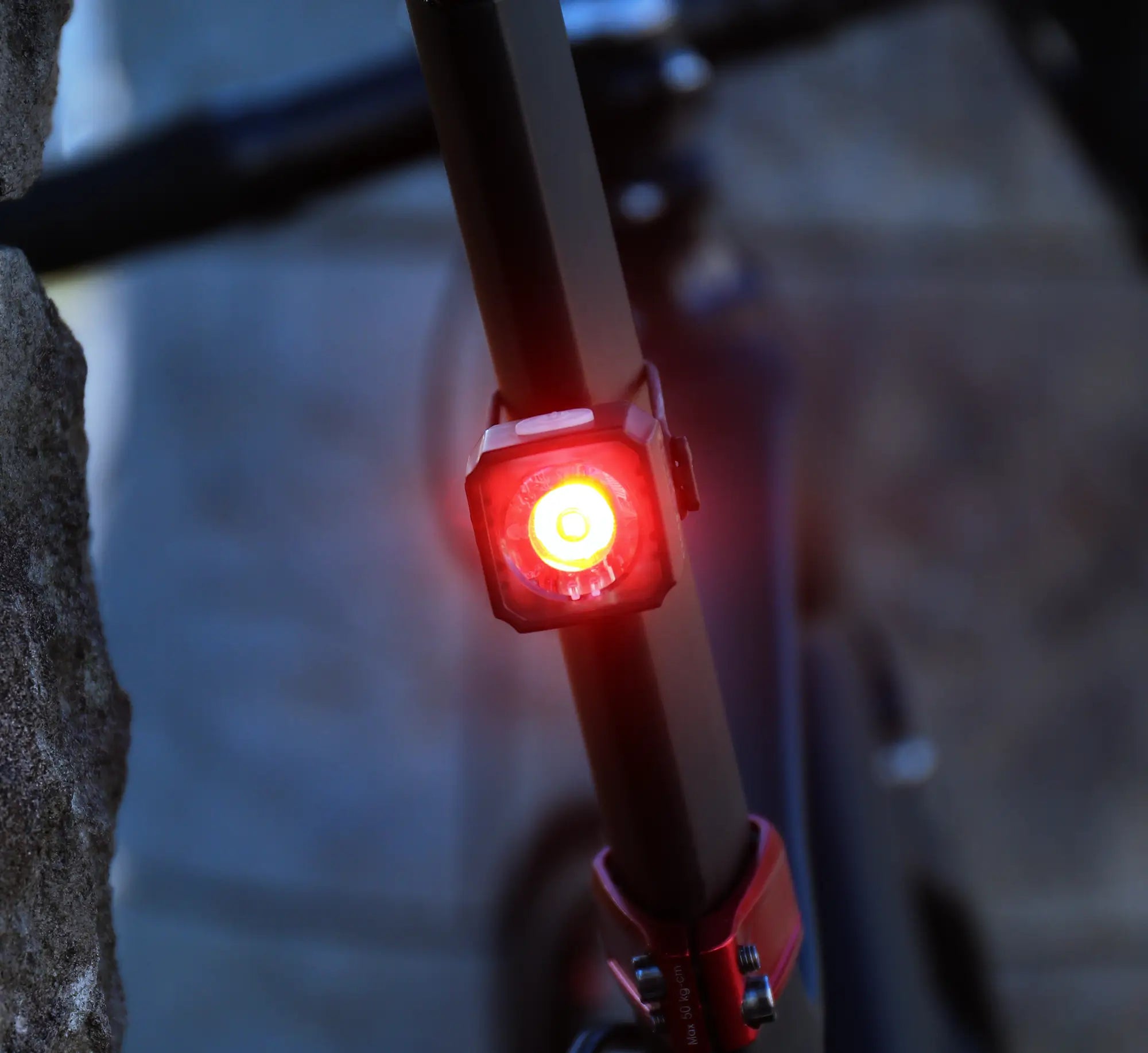 A close-up of the CATEYE AMPP400S & NANO60 red LED bike light glowing brightly on a seat post, with a stone wall and blurred bike frame in the background.