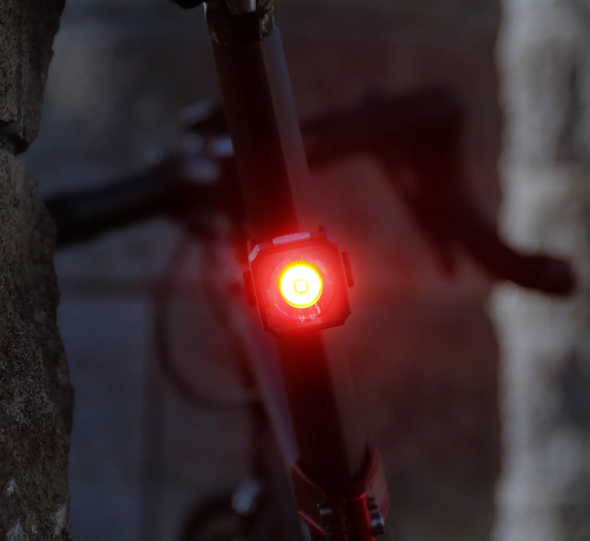 A close-up of the CATEYE NANO60, a bright red USB-C rechargeable bike light attached to a seat post, with a blurred bike and stone wall in the background, highlights its enhanced visibility in dusk or low-light conditions.