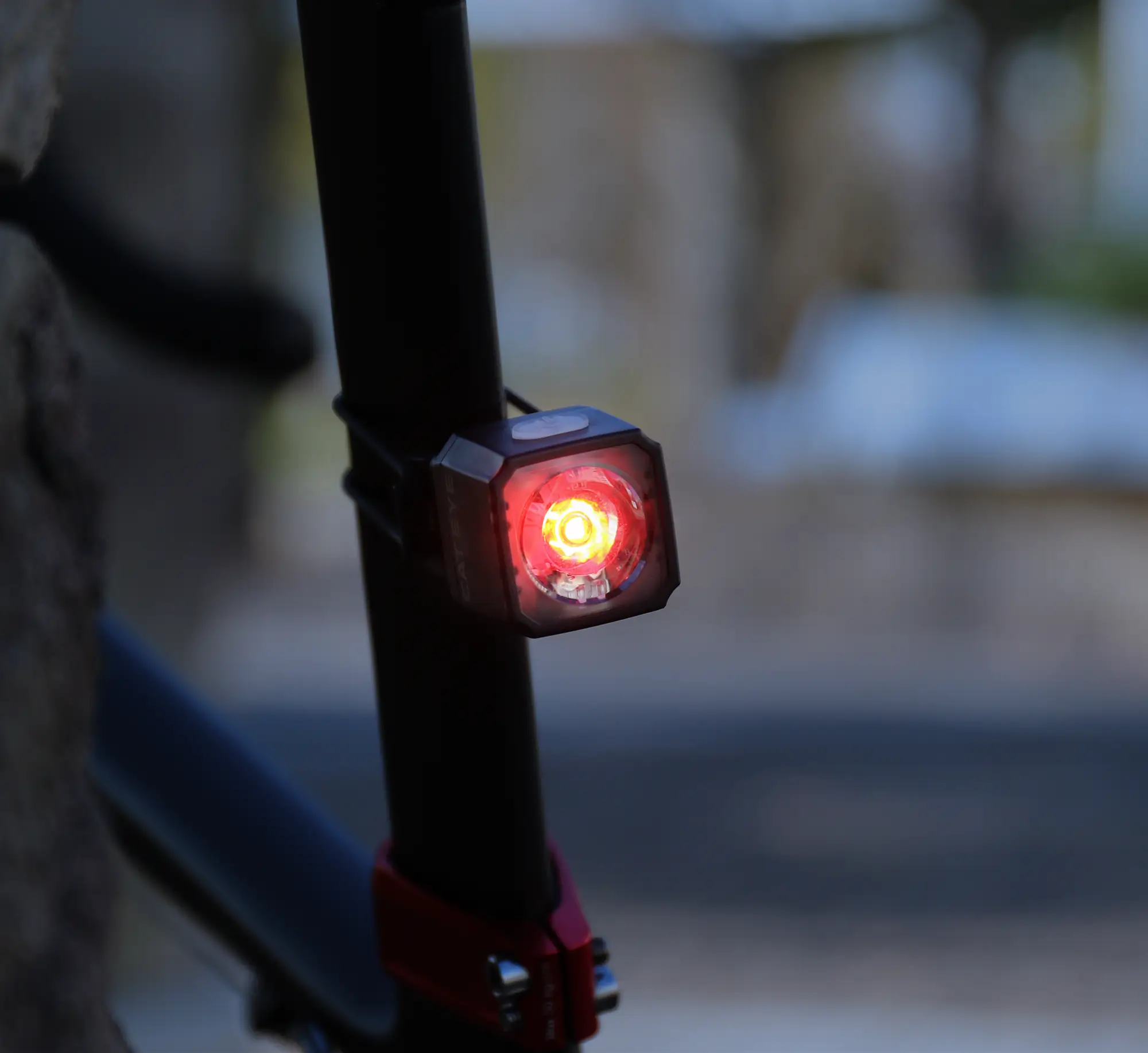 A close-up of the CATEYE NANO60, a red USB-C rechargeable bike light, attached to a seat post and glowing brightly for enhanced daytime visibility in a dim outdoor setting.