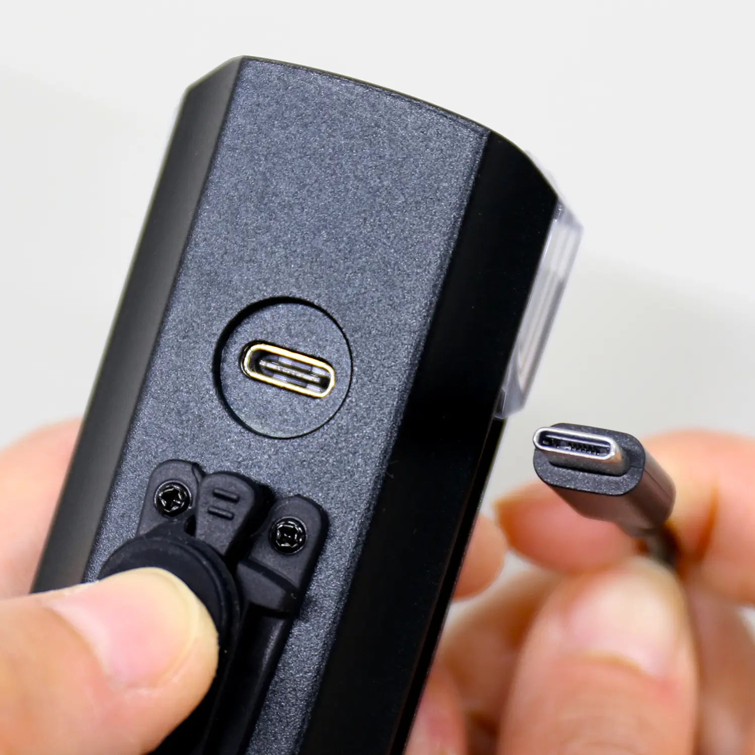 A close-up of hands holding a CATEYE AMPP1300 light and a USB-C cable, aligning the cable with the AMPP1300’s USB-C port for charging or data transfer.