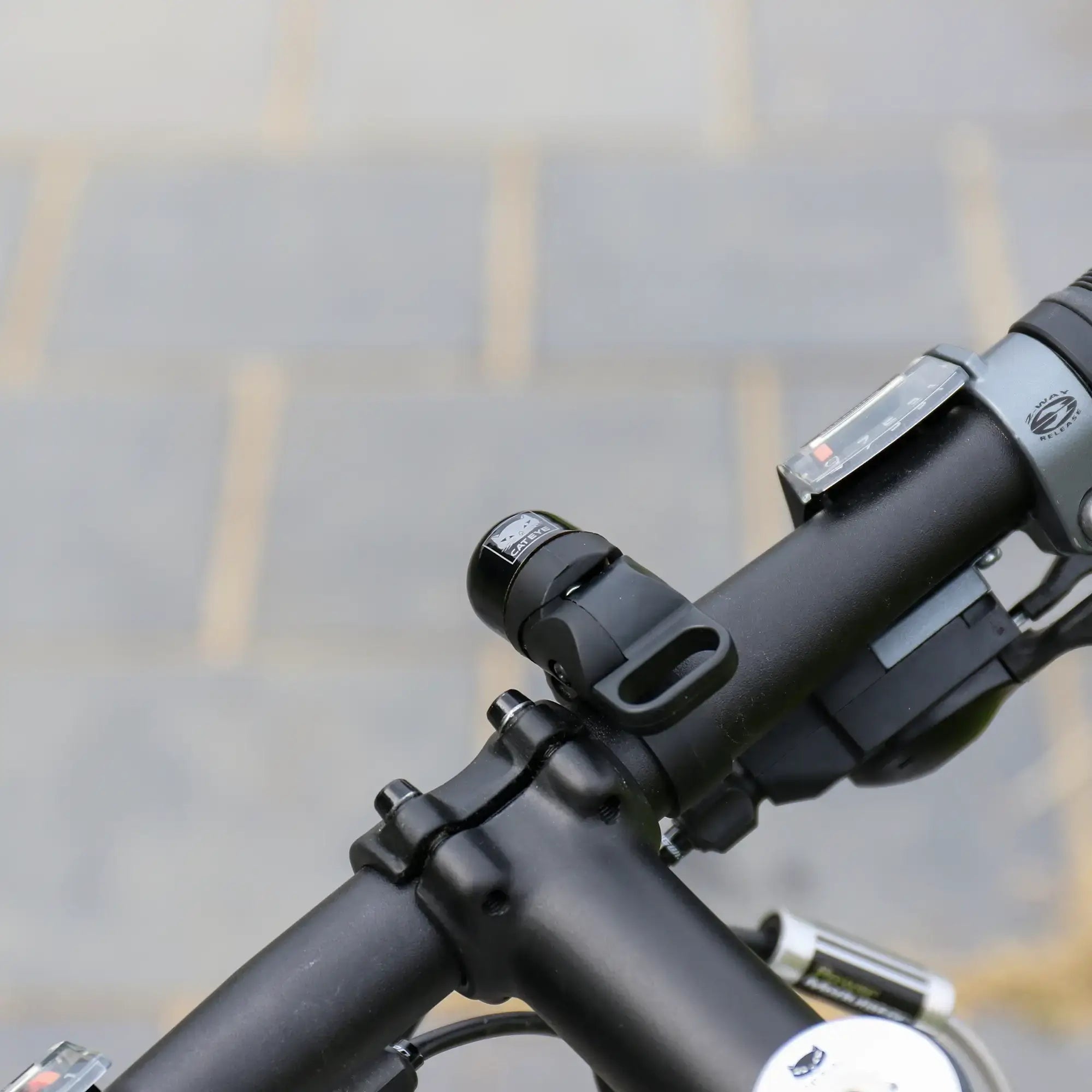 Close-up of a CATEYE OH-1500 NIDO Bell mounted on a black adjustable handlebar, showing the stem, gear shifter, part of the brake lever, and blurred tiled pavement in the background.