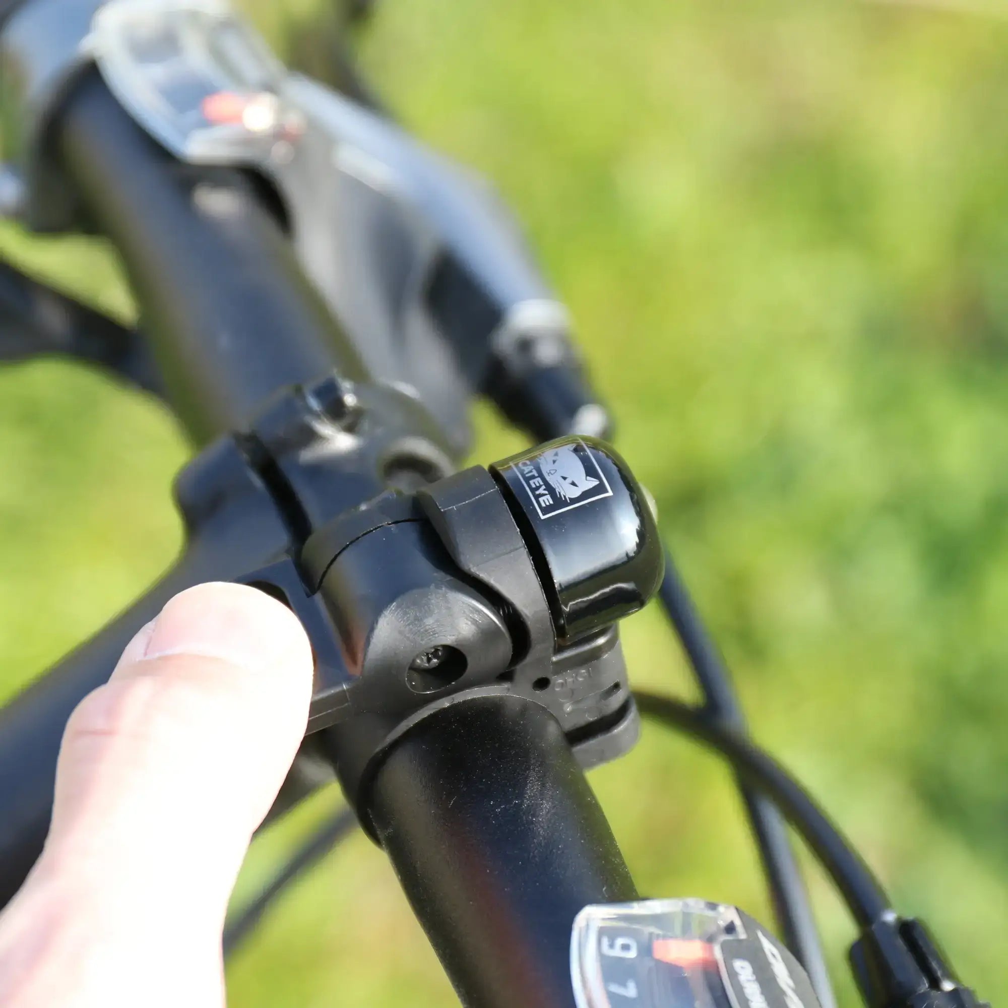 A close-up shows a person’s hand adjusting a CATEYE OH-1500 NIDO Bell with a quick-release lever on a bicycle handlebar, with blurred greenery in the background.