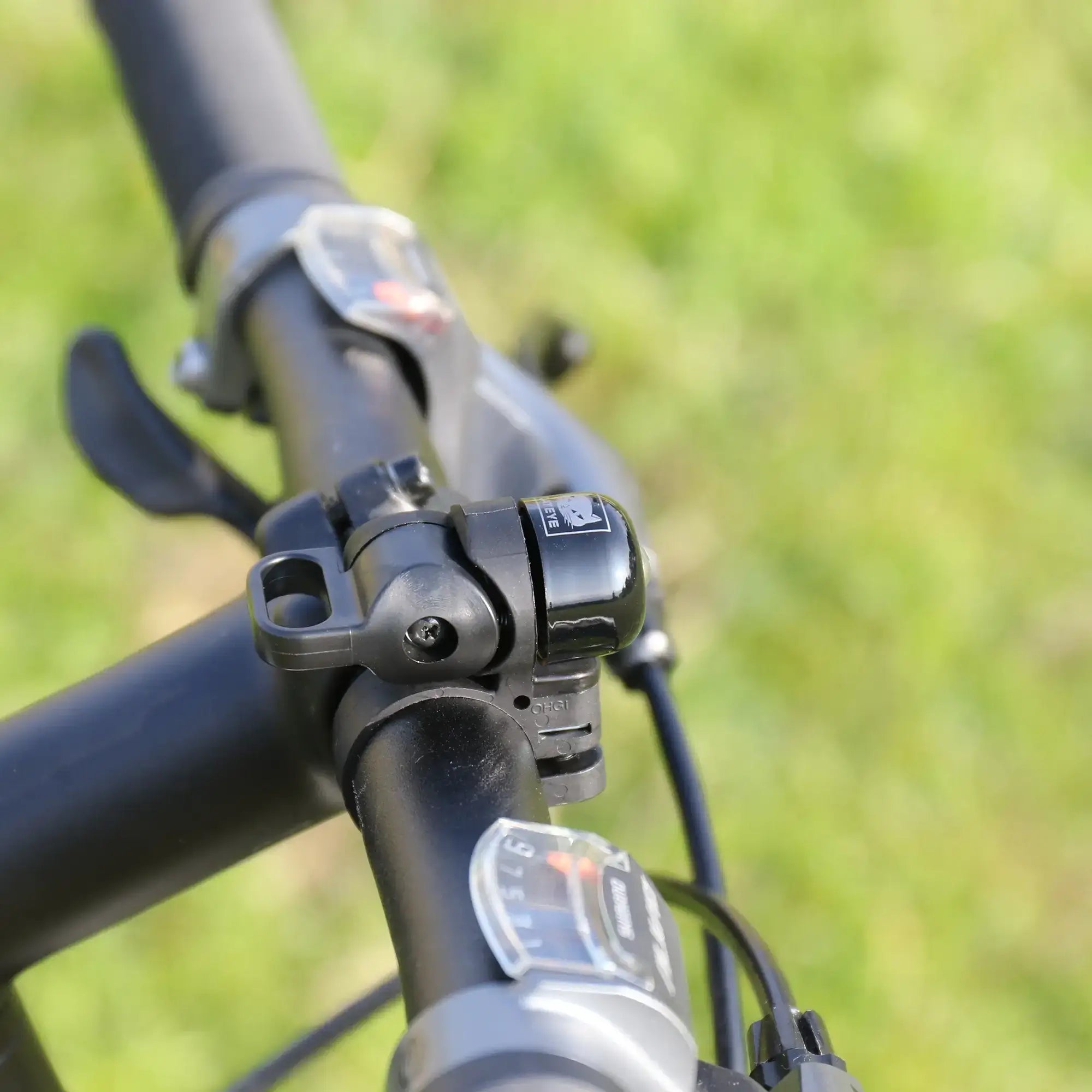Close-up of a CATEYE OH-1500 NIDO Bell mounted on a bicycle handlebar, with gear shifters and brake levers, set against a blurred green grass background.