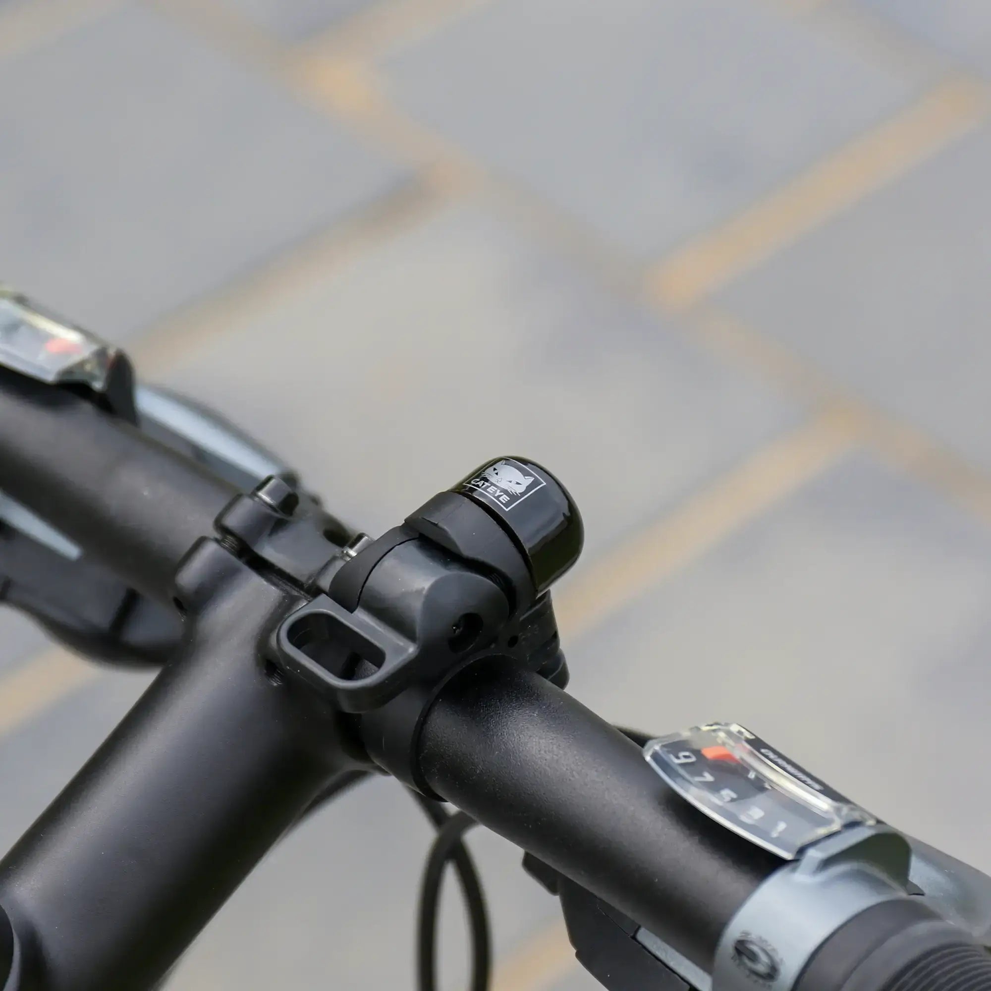 Close-up of a black adjustable handlebar featuring the CATEYE OH-1500 NIDO Bell and visible gear shifters on both sides, set against a blurred paved background.