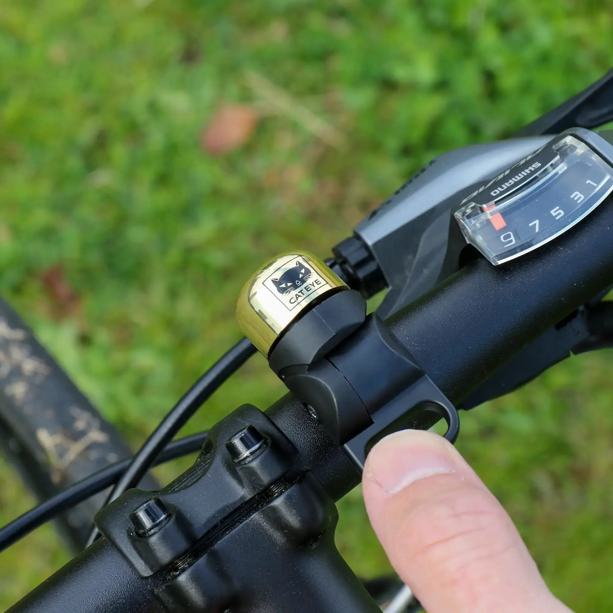 A close-up of a person pressing the CATEYE OH-1500 NIDO Bell, featuring a cat face design on a black adjustable handlebar beside a gear shifter, with grass in the background.