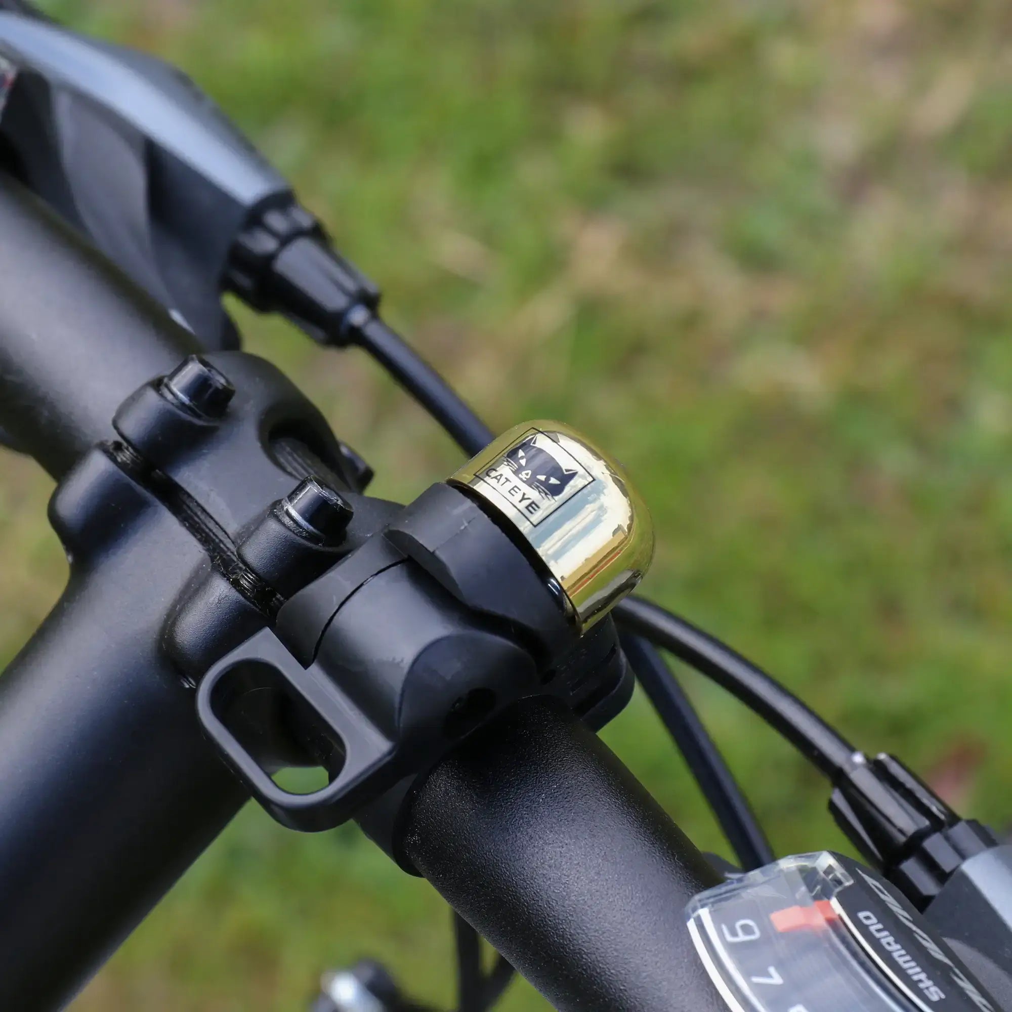 Close-up of a CATEYE OH-1500 NIDO Bell attached to a bicycle handlebar, secured by a gold padlock on an adjustable clamp. The bell is prominent, with a softly blurred grassy background.