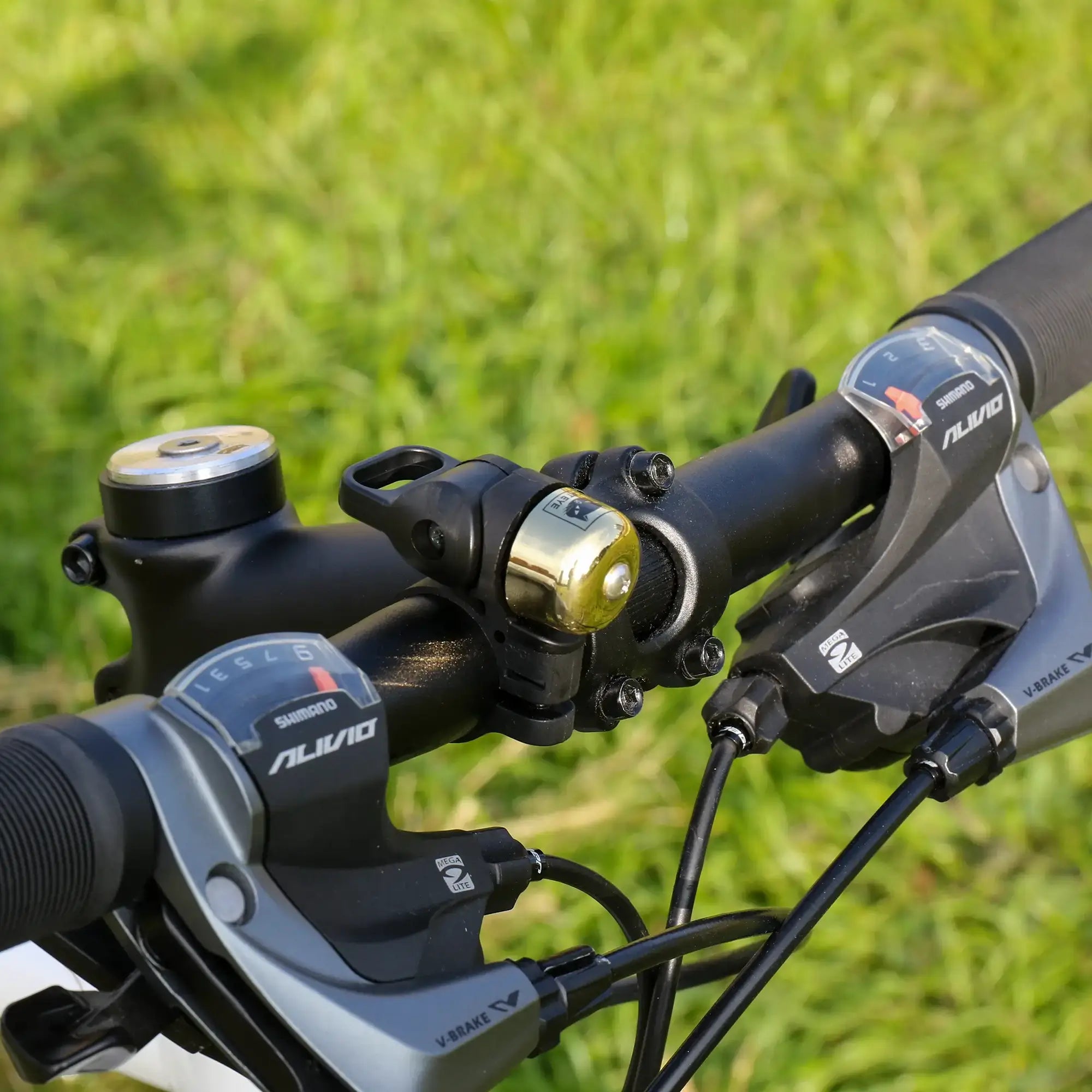 Close-up of an adjustable handlebar with Shimano Alivio shifters, black grips, and a compact metallic CATEYE OH-1500 NIDO Bell, plus cables; green grass is blurred in the background.