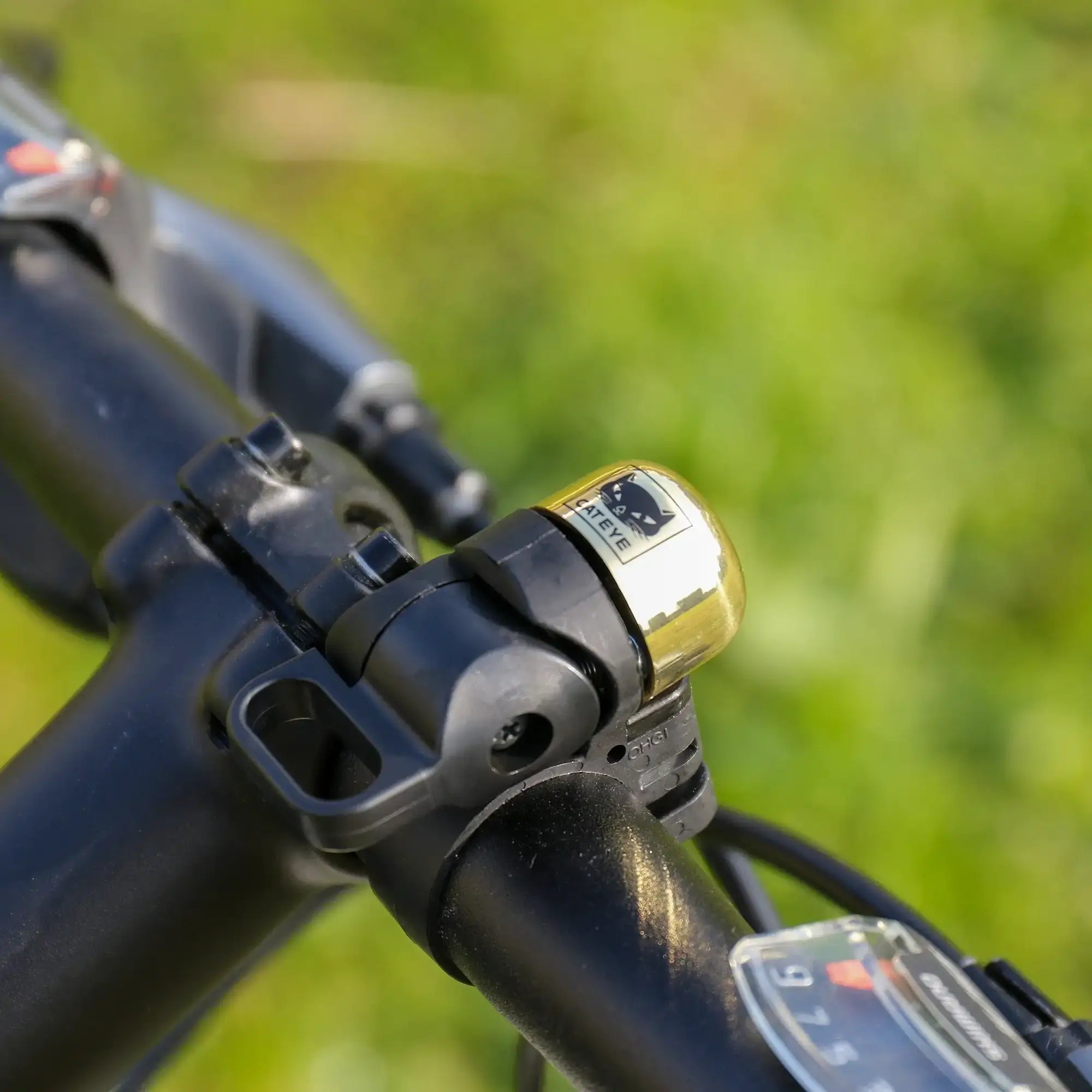 Close-up of an adjustable handlebar featuring a shiny gold and black CATEYE OH-1500 NIDO Bell, labeled CATEYE, with a blurred green outdoor background.