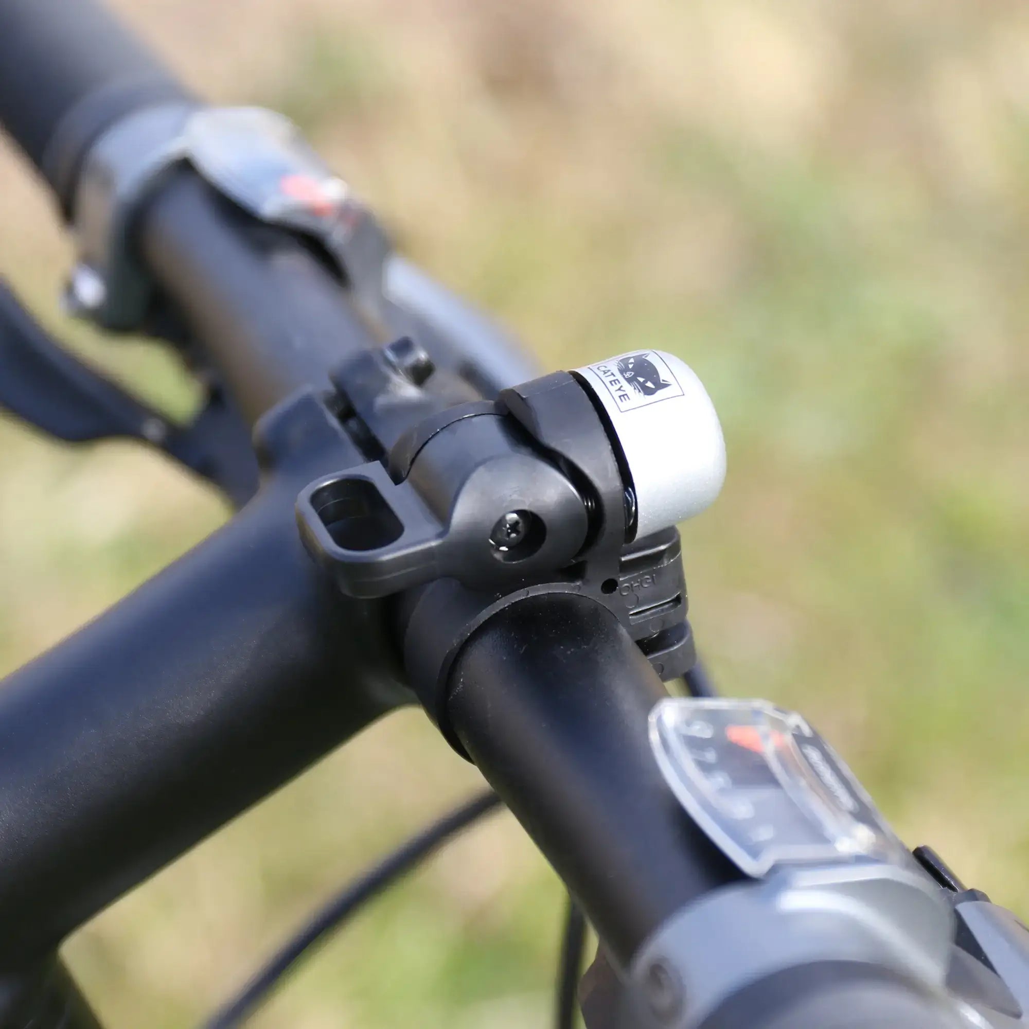 A compact silver and black CATEYE OH-1500 NIDO Bell is mounted on a bike’s adjustable handlebar, with an outdoor background softly blurred.