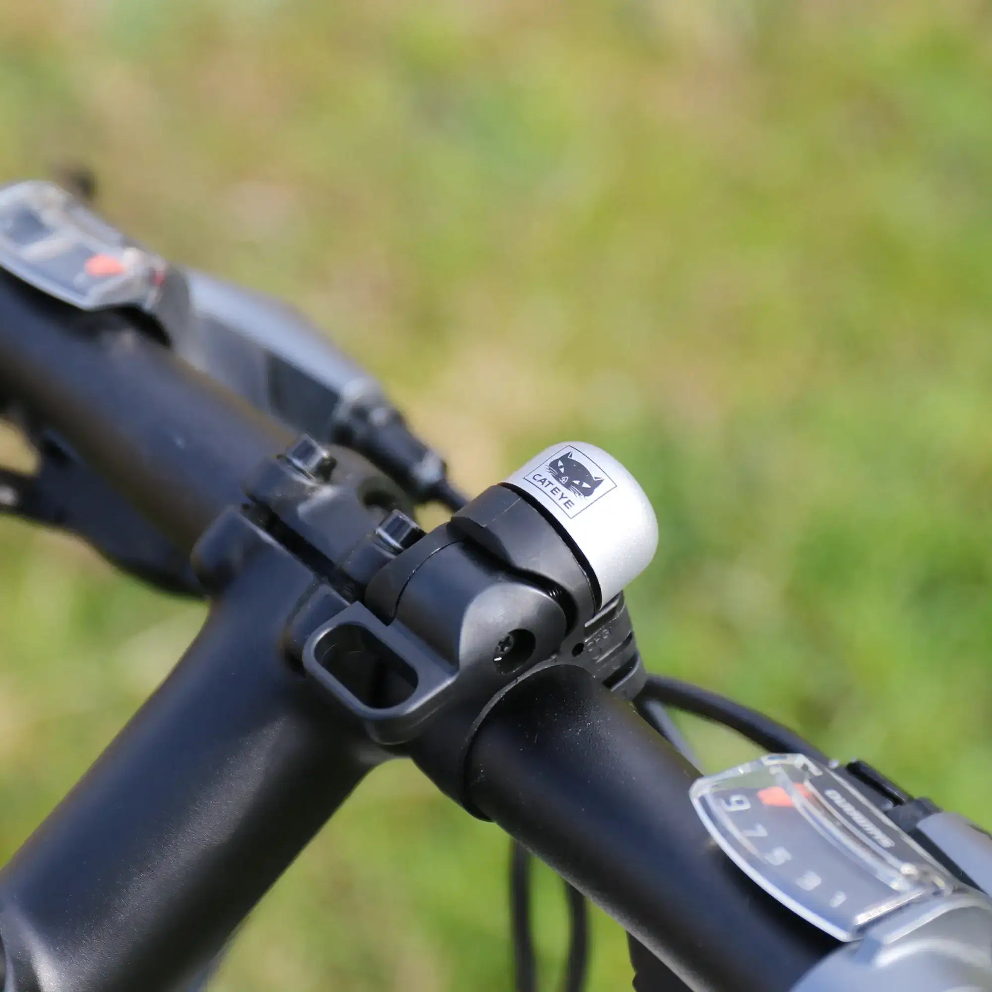Close-up of a bicycle handlebar featuring the CATEYE OH-1500 NIDO Bell in silver and black, with gear shifters on each side. The adjustable handlebar stands out against a blurred green grass background.