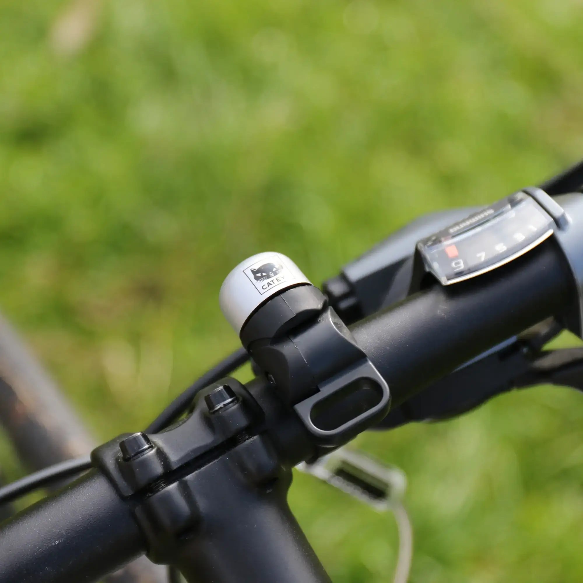 Close-up of the CATEYE OH-1500 NIDO Bell—a small silver and black bicycle bell with a cat logo—mounted beside a gear shifter on an adjustable handlebar, set against blurred green grass.