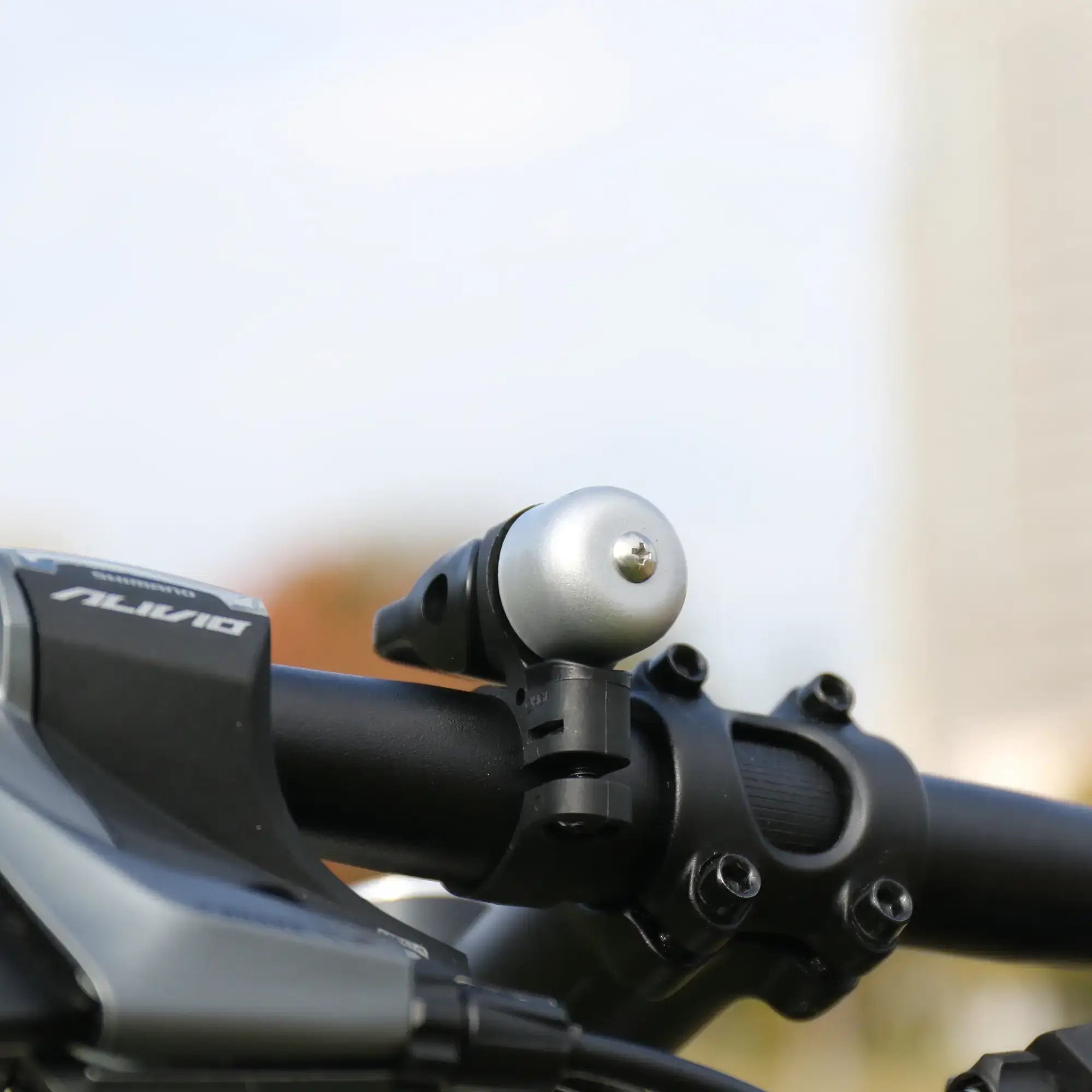 A close-up of the CATEYE OH-1500 NIDO Bell mounted on an adjustable bike handlebar, with part of the gear shifter in view and a blurred outdoor background.