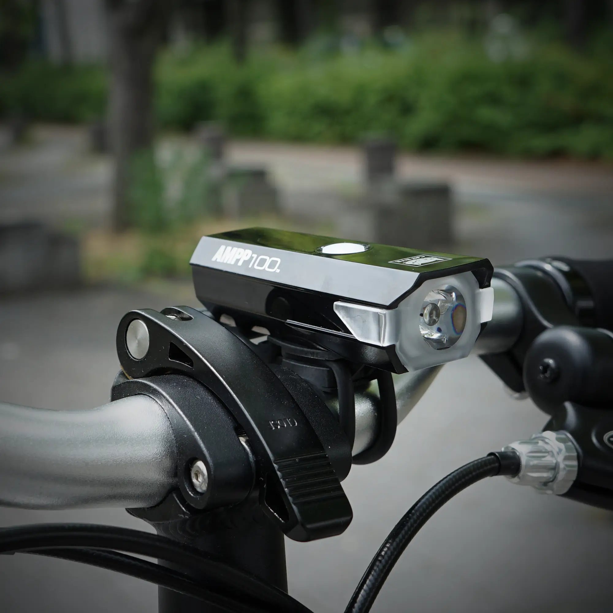 A close-up of a CATEYE AMPP100 & ORB set mounted on a black bike handlebar, featuring OptiCube™ lens technology and showcasing its sleek design against an urban outdoor backdrop.