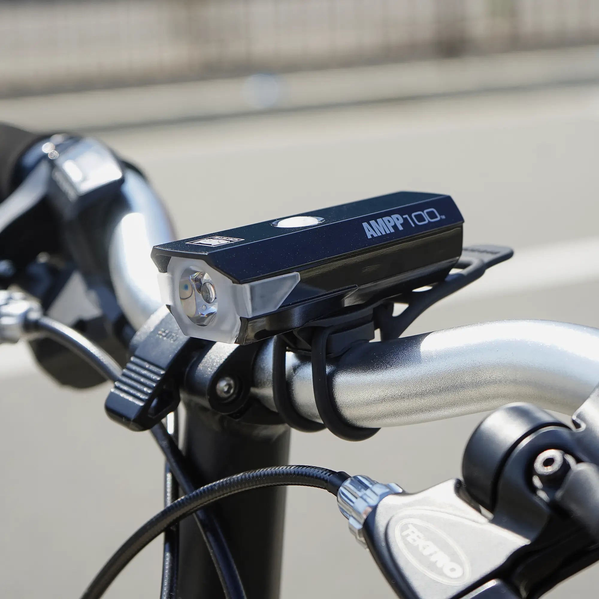 A black CATEYE AMPP100 & ORB bike headlight with OptiCube™ lens technology is mounted on a bicycle’s handlebars, shown in daylight with a blurred street background.