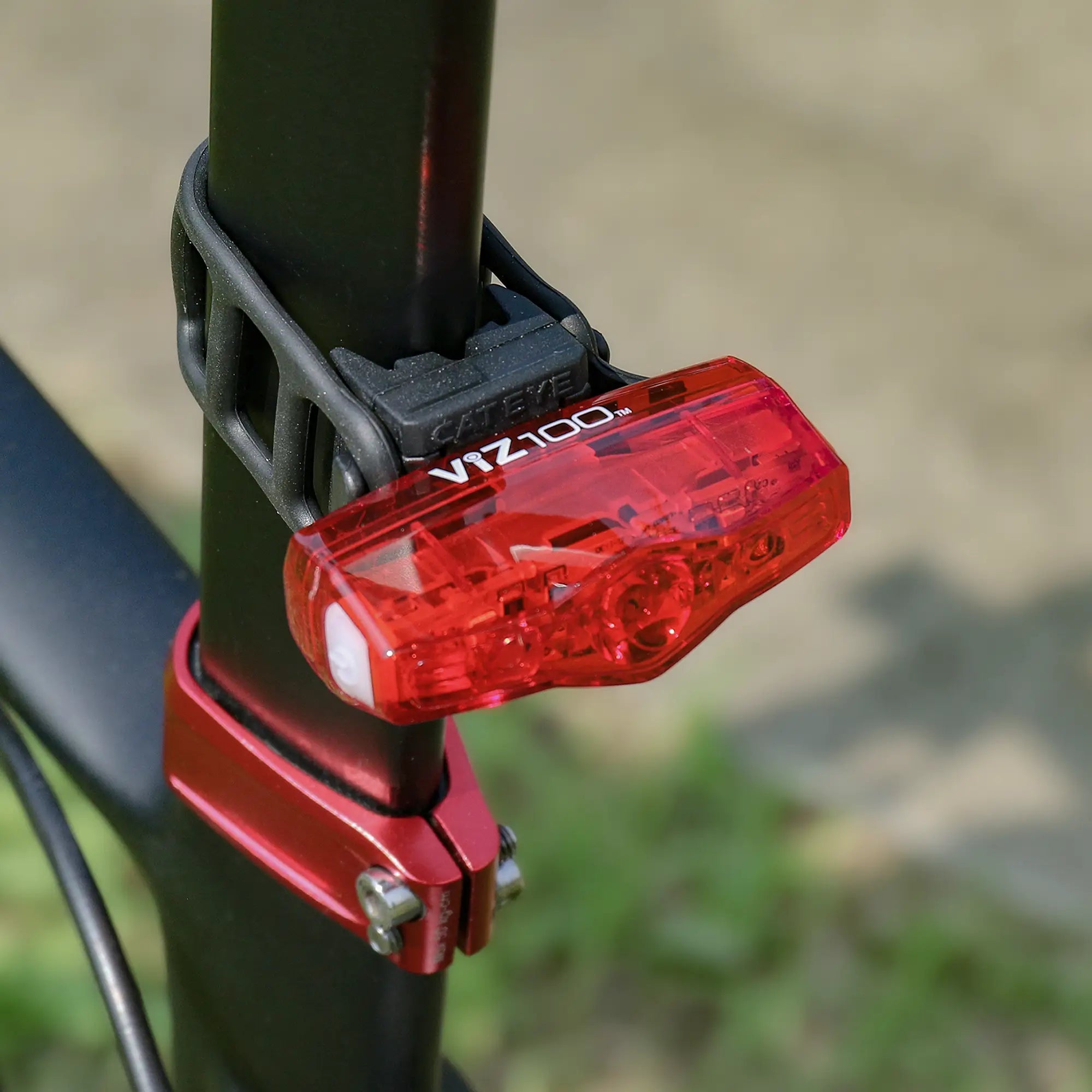 The CATEYE ViZ100 red bike tail light, part of the AMPP200 & ViZ100 set, is secured to a black seat post with a rubber strap. USB rechargeable, it stands out against blurred grass and pavement in the background.