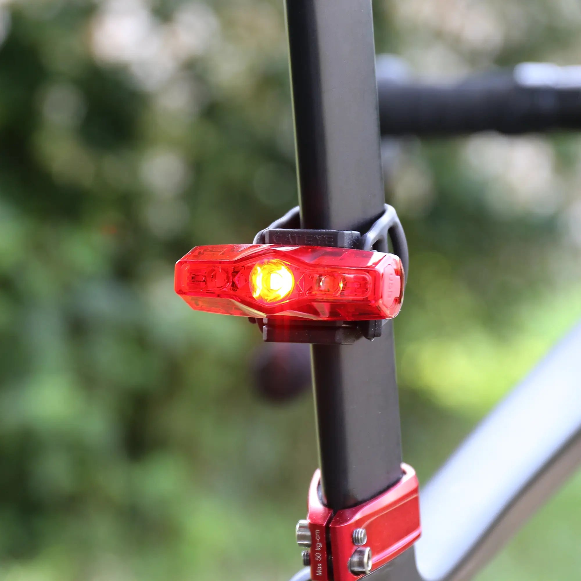The CATEYE AMPP200 & ViZ100 features a red LED tail light with a yellow center glowing brightly on the seat post. Its USB rechargeable design offers convenience, set against a backdrop of blurred greenery.