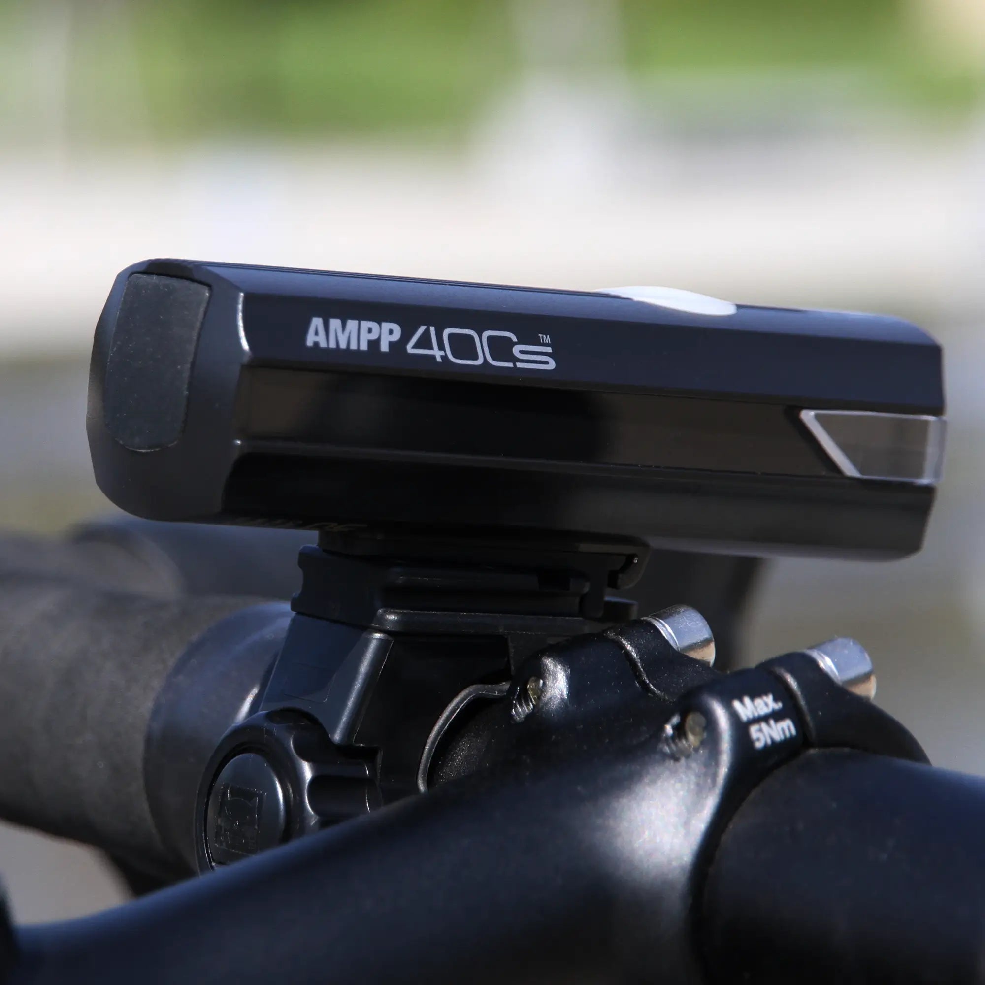 Close-up of a black CATEYE AMPP400S & ViZ100 bicycle headlight with OptiCube™ lens technology, mounted on handlebars. Handlebar clamp and adjustment bolts are visible, with a blurred background.