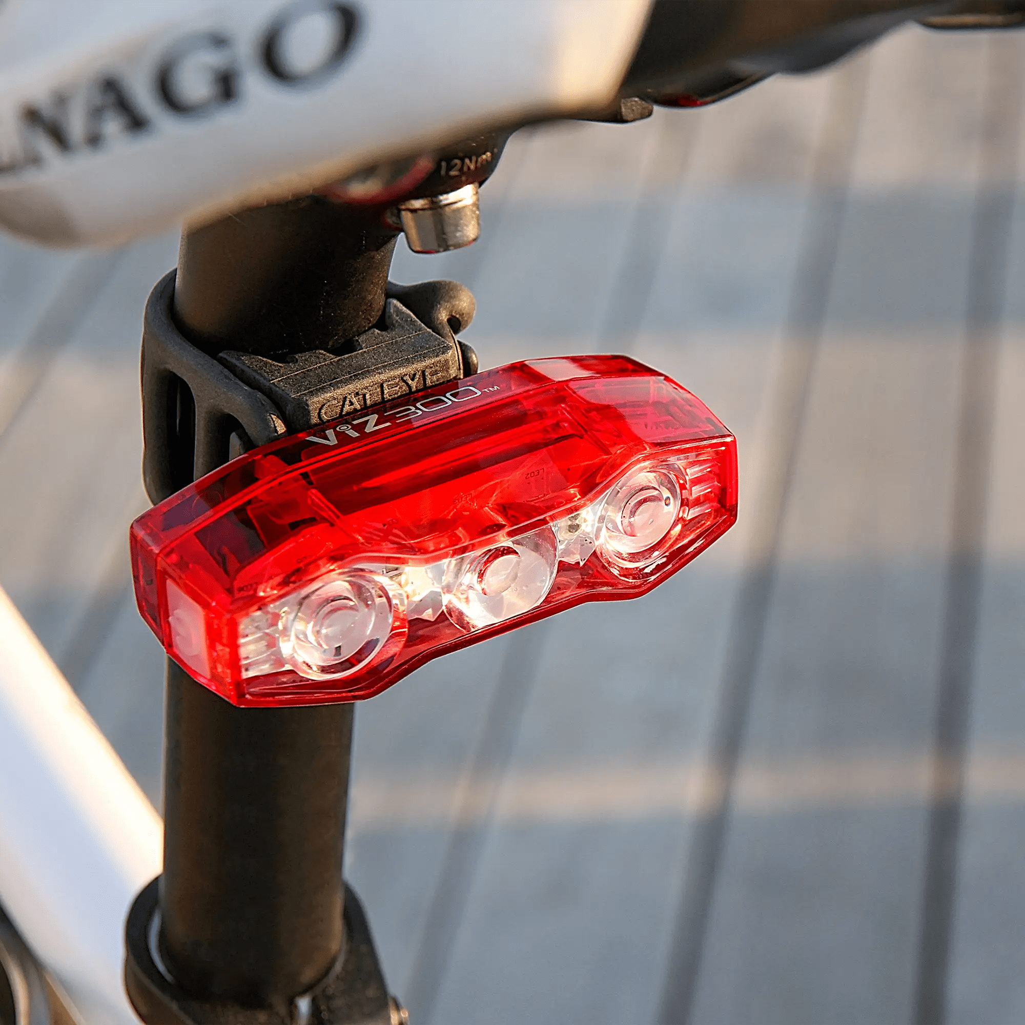 A CATEYE ViZ300 (USB-C) red bicycle tail light is mounted on a bike seat post, with a blurred wooden background. Its clear cover exposes inner components and features OptiCube™ lens technology for enhanced visibility.