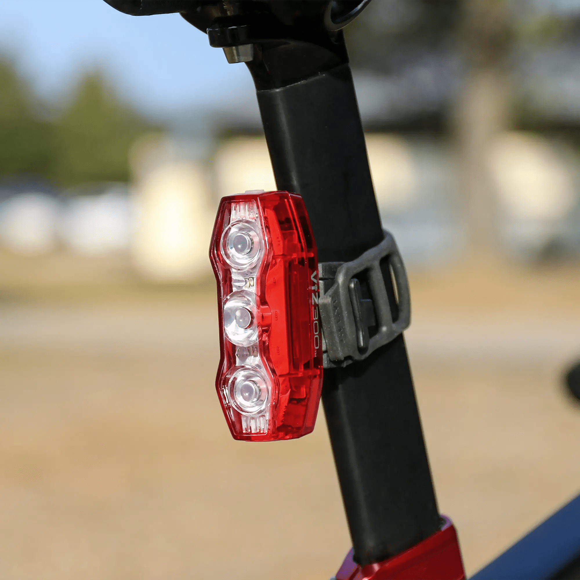 A CATEYE ViZ300 (USB-C) LED tail light with OptiCube™ lens, in red and white, is mounted to a bike seat post using a black rubber strap. The outdoor background appears blurred. Also shown: AMPP900 front light.