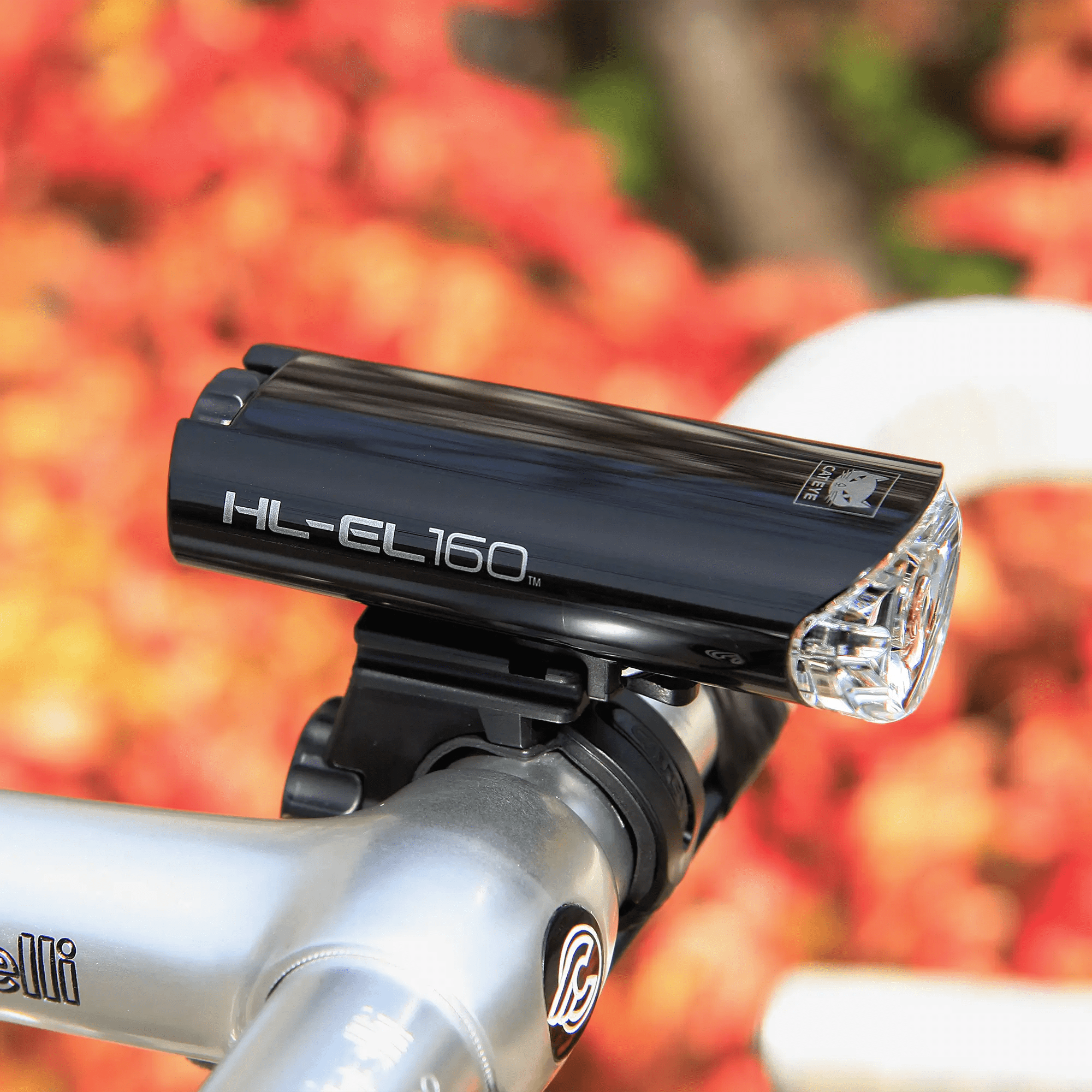 A black, waterproof CATEYE HL-EL160 & OMNI 5 headlight is mounted on silver bicycle handlebars, providing 360° visibility against a softly blurred backdrop of red and orange foliage.