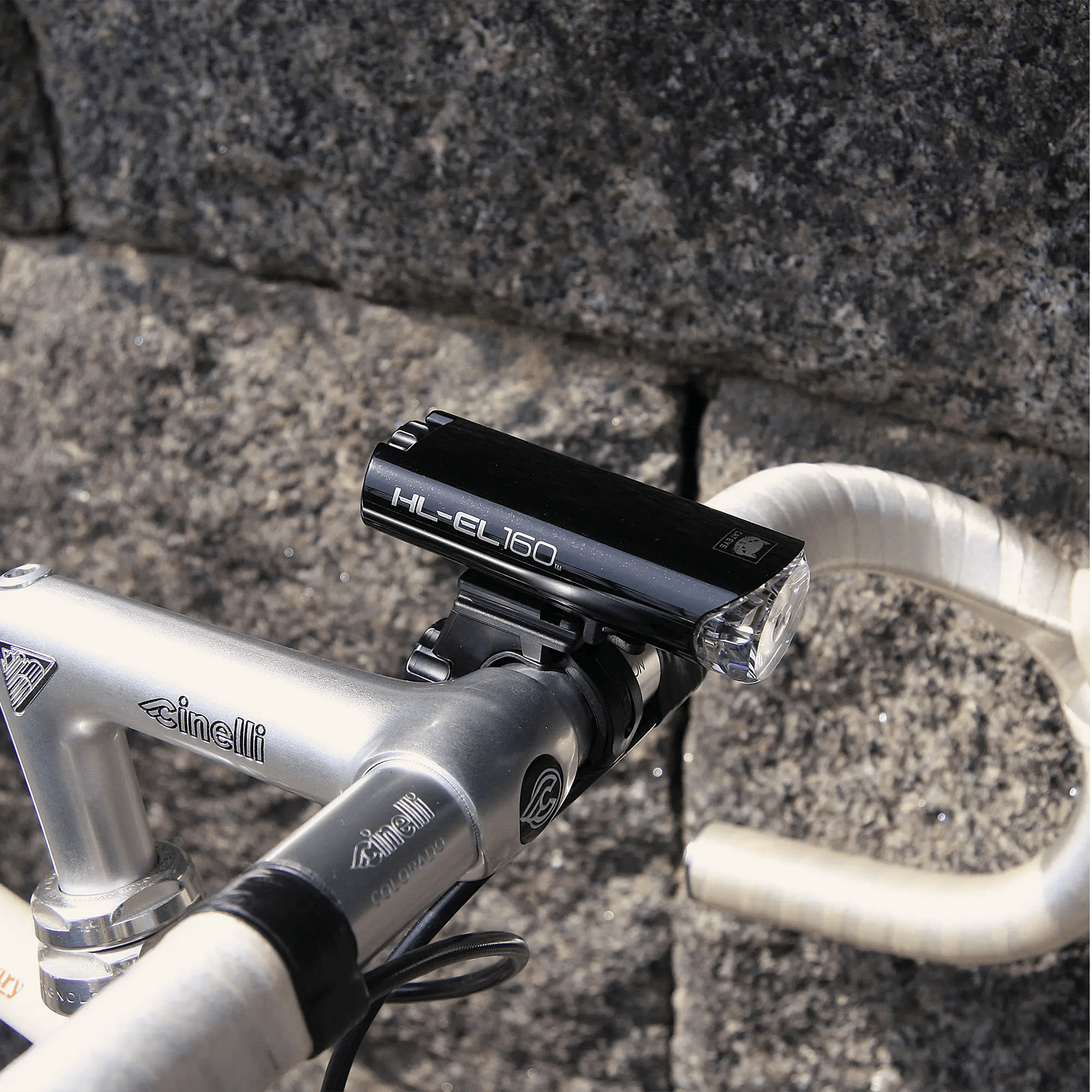 A black CATEYE HL-EL160 & OMNI 5 waterproof headlight is mounted on a silver Cinelli bike’s handlebars in front of a stone wall, providing 360° visibility for safer rides.