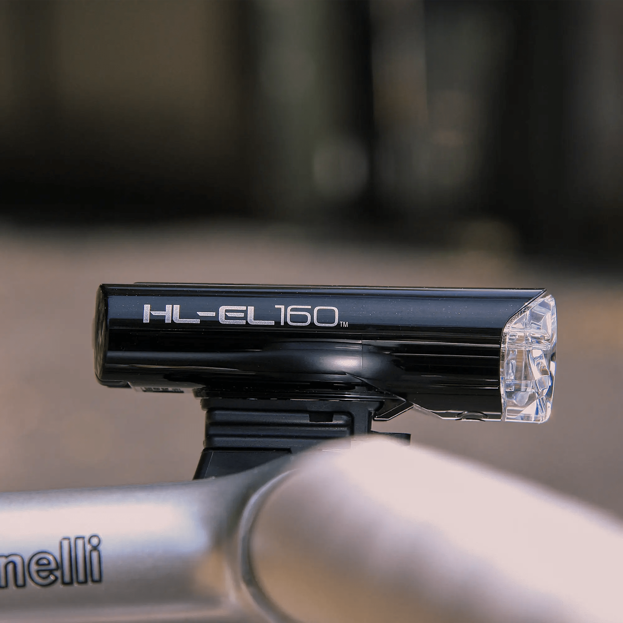 A close-up of the black CATEYE Hl-El160 & OMNI 5 waterproof headlight mounted on a silver bike handlebar, with a blurred outdoor background.