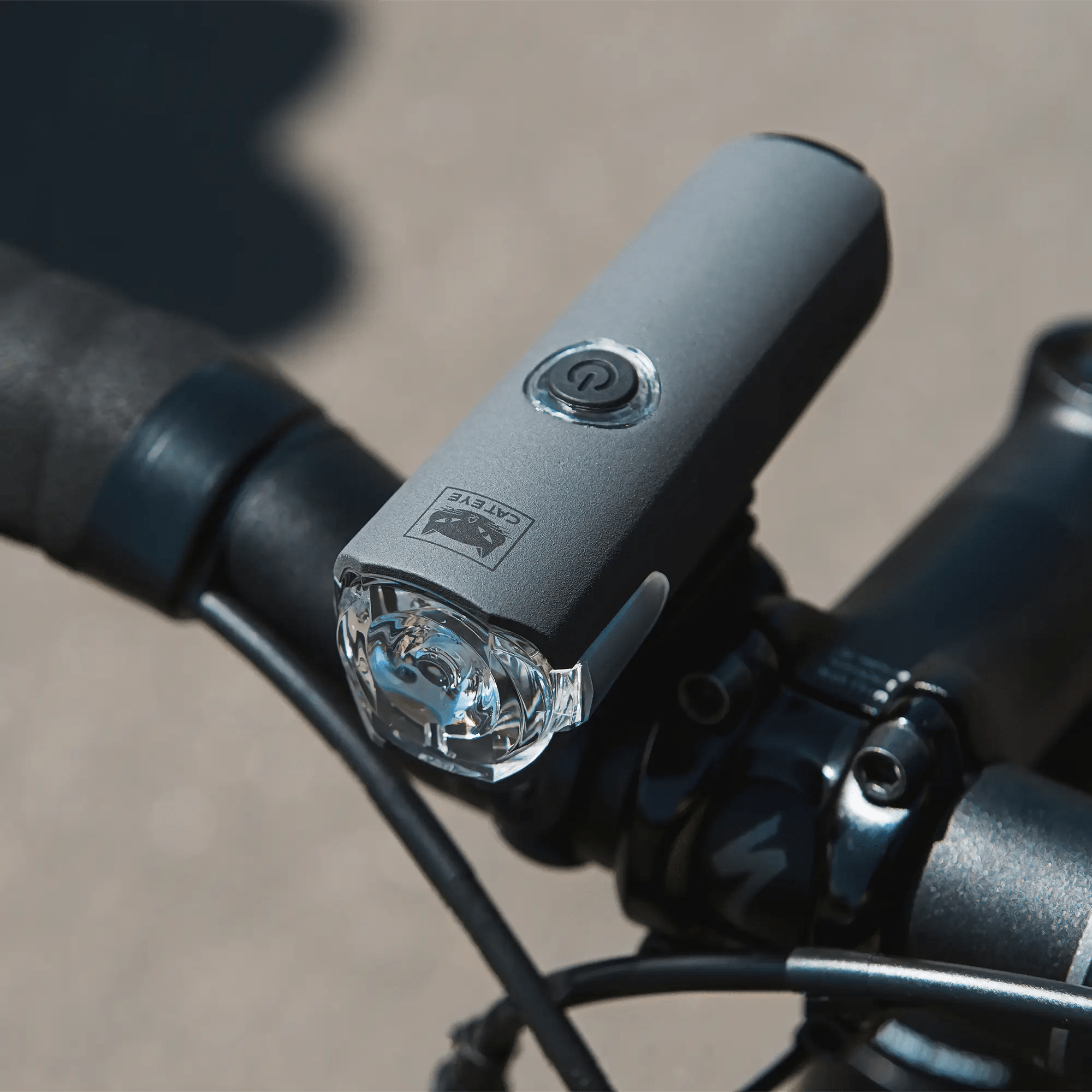 A close-up of a black bicycle handlebar with a mounted CATEYE SYNC Core & SYNC Kinetic front light featuring a power button, clear lens, and smartphone app control as sunlight casts shadows on the pavement.