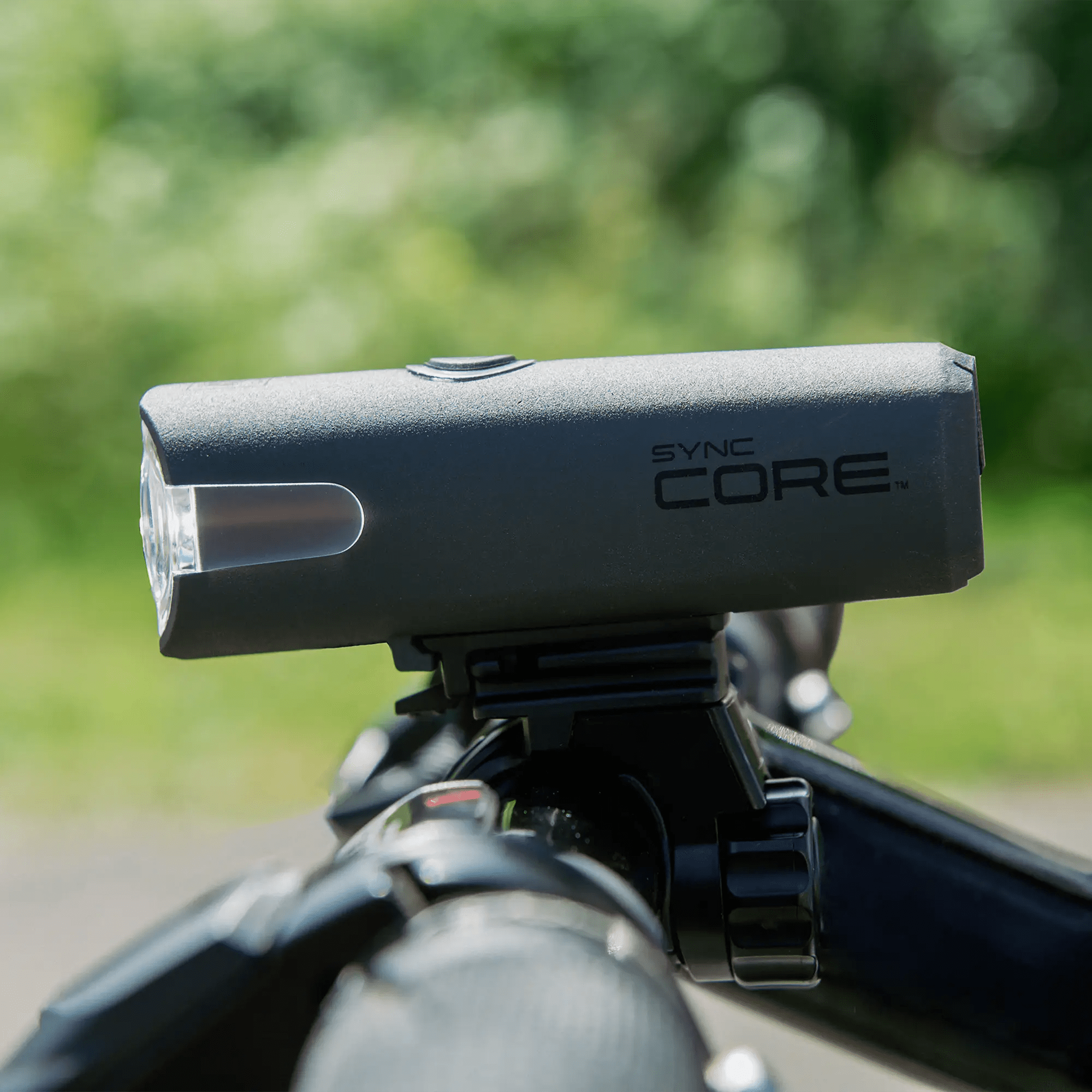 A close-up of the CATEYE SYNC Core & SYNC Kinetic bicycle headlights, mounted on a handlebar, showcasing innovative lighting technology against a blurred green background.