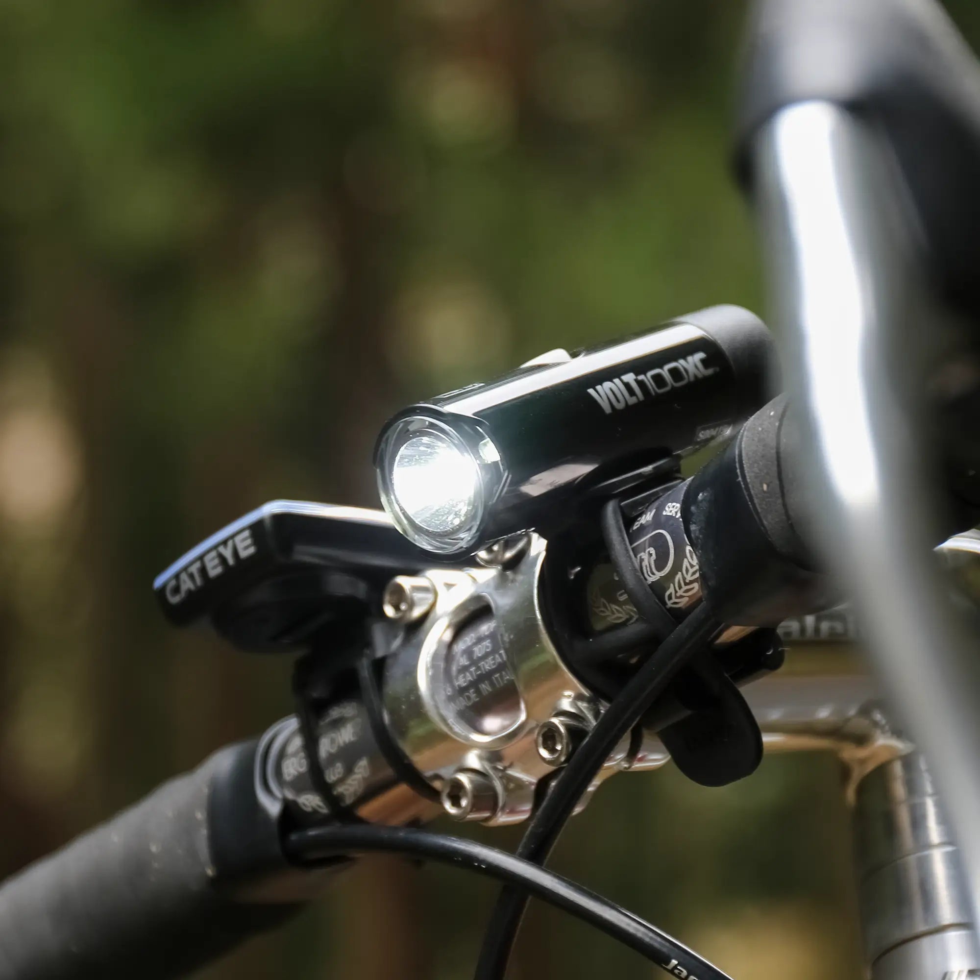 Close-up of a CATEYE VOLT100XC & ORB handlebar combo—USB rechargeable headlight and cycling computer—mounted on durable aluminum, illuminated with a blurred green background.